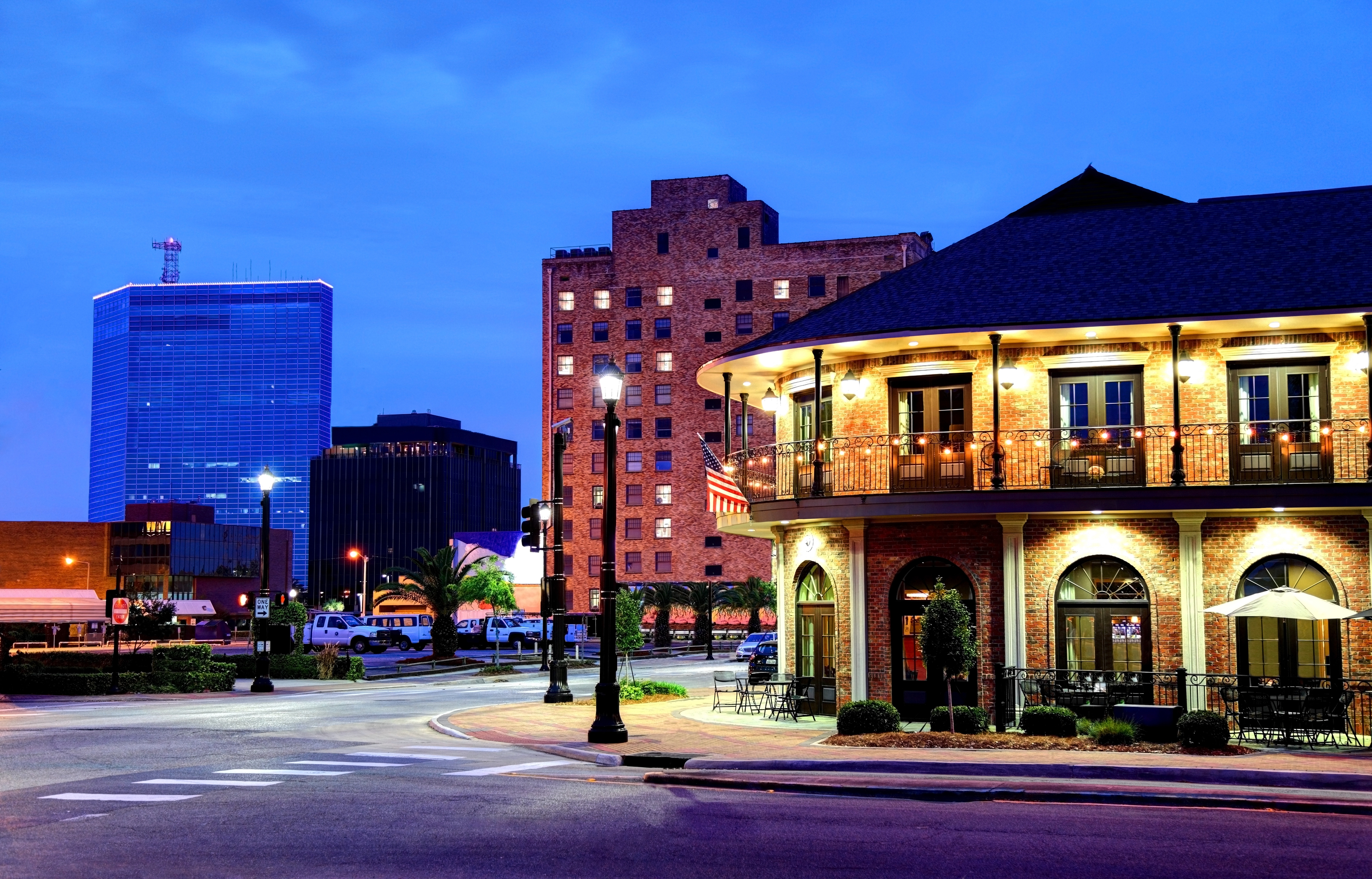 A charming evening view of downtown Lake Charles, Louisiana, featuring a well-lit historic brick building with a balcony and modern skyscrapers glowing under a deep blue sky.