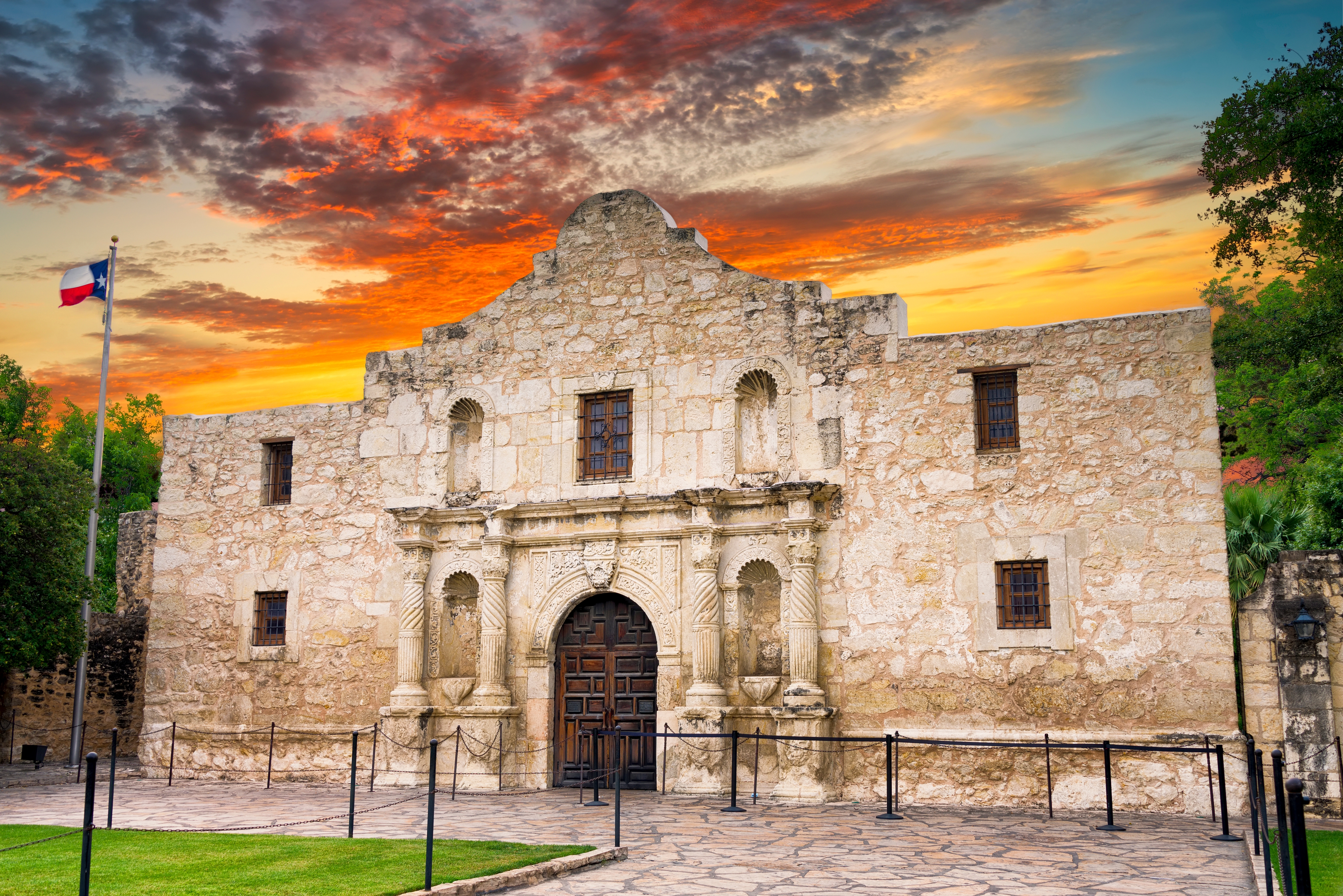 Historic Alamo mission in San Antonio, Texas, captured at sunset with vibrant skies and the Texas flag waving beside the iconic stone facade.