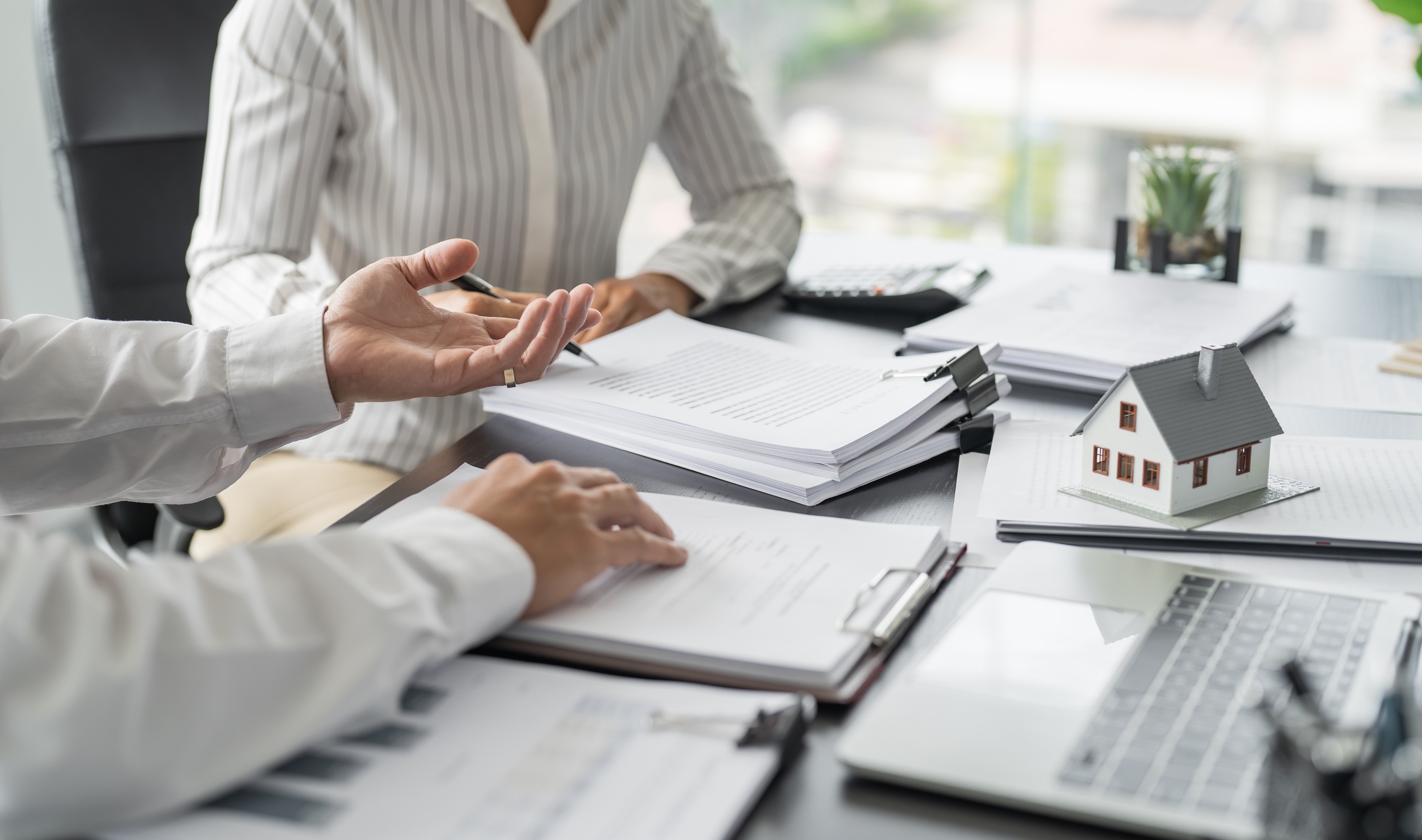 Close-up of a real estate meeting with paperwork, a miniature house model, and two professionals discussing documents at a modern office desk.