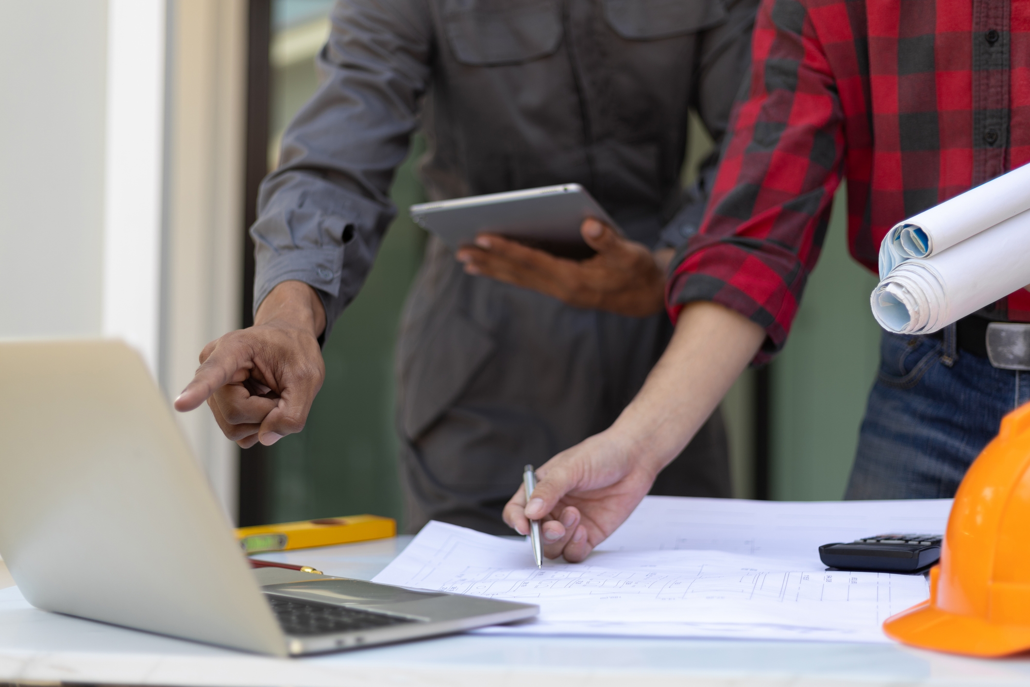 Close-up of two real estate professionals reviewing blueprints and data on a laptop at a worksite, with one holding rolled plans and the other pointing at the screen.