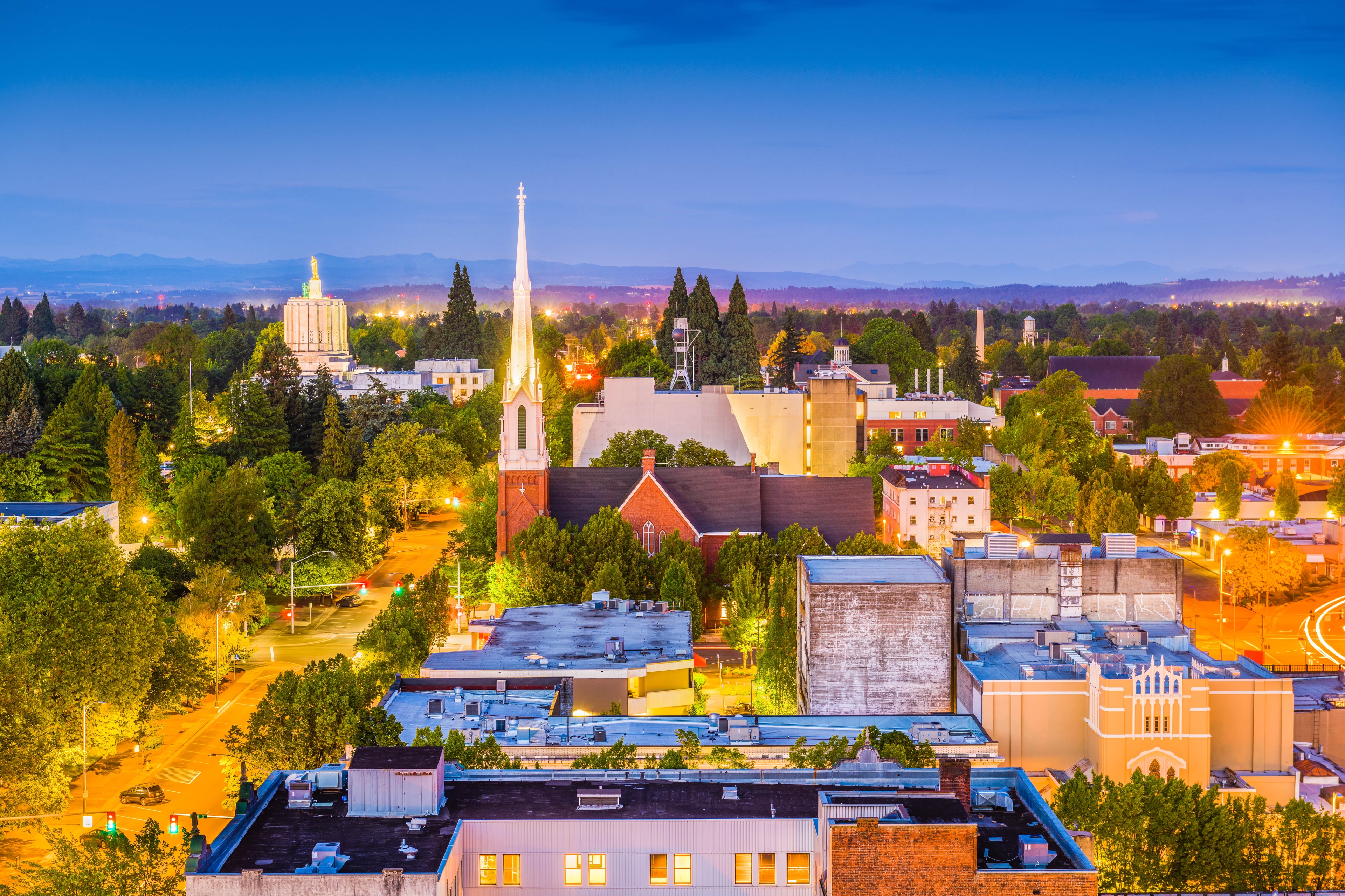 Evening view of downtown Salem, Oregon, with illuminated streets, historic churches, and the Oregon State Capitol visible among green trees and distant hills.