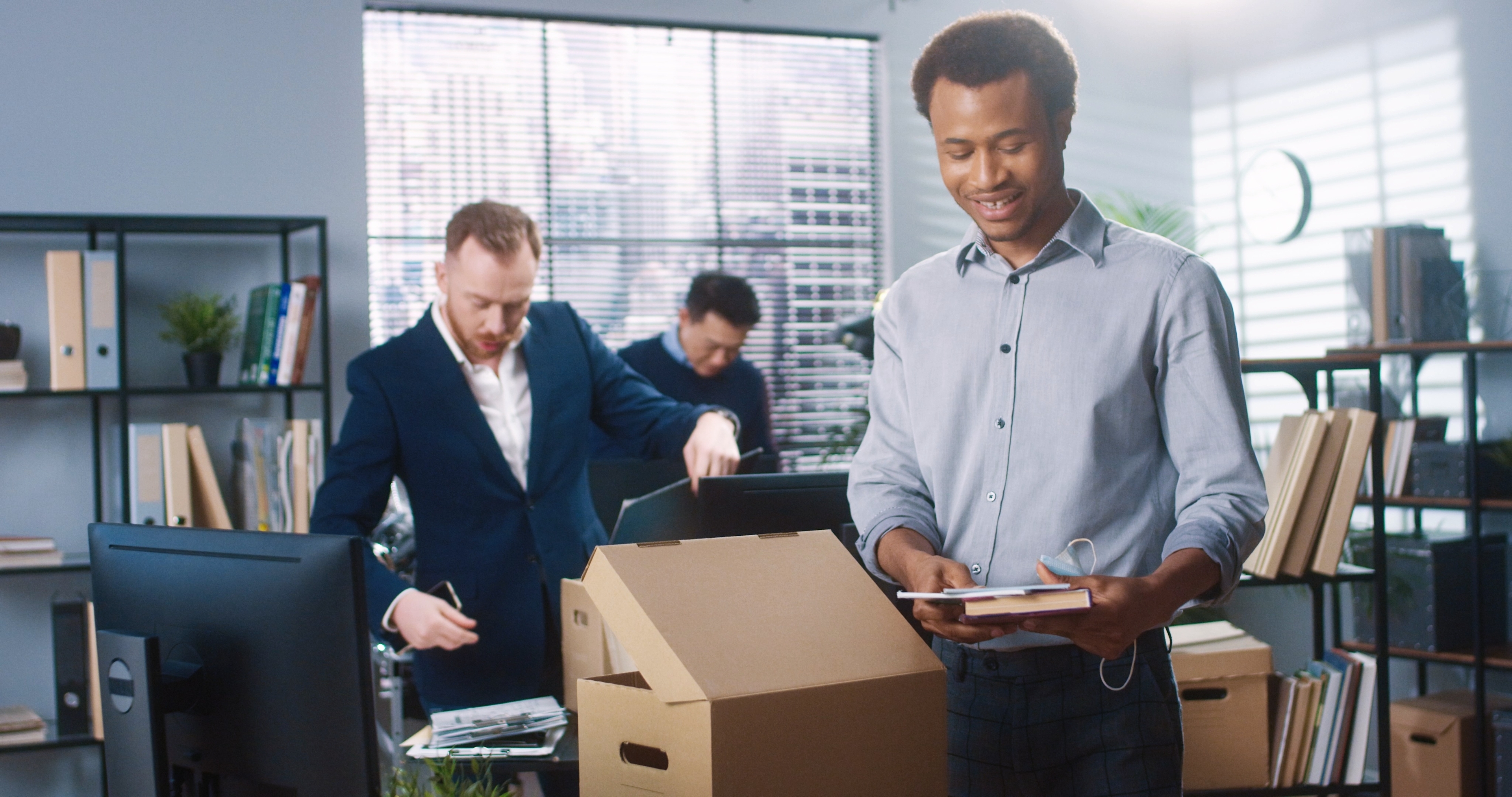 Three colleagues working in a modern office environment, with one unpacking books from a box while others organize papers and use a computer. The room includes bookshelves, desks, and a bright window with blinds.