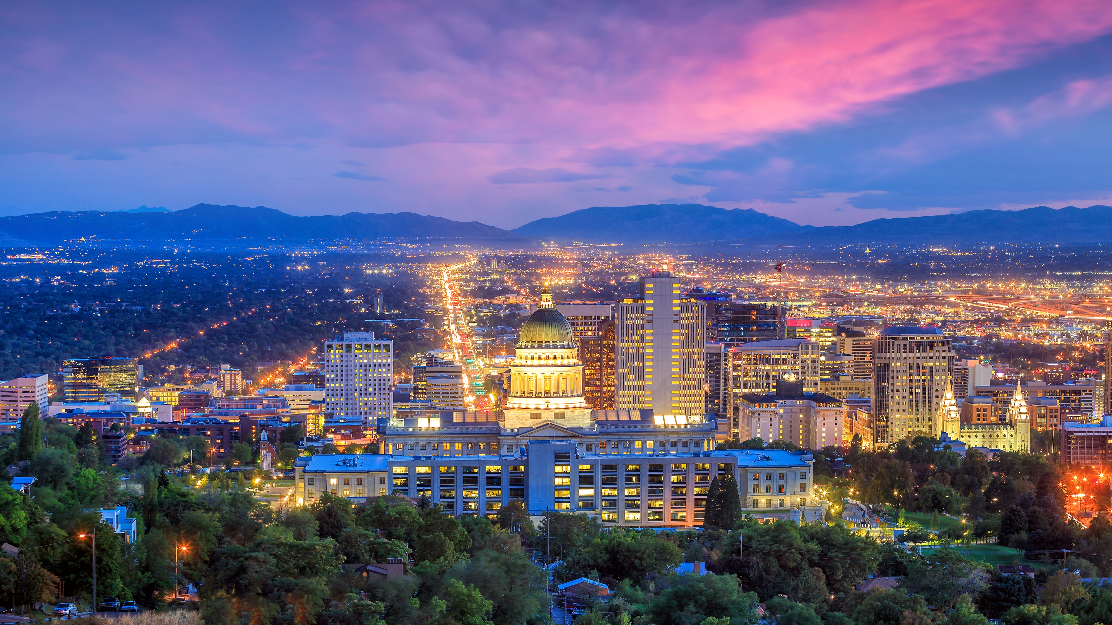 Evening view of downtown Salt Lake City, Utah, with the illuminated Utah State Capitol building in the foreground, a glowing skyline behind it, and a vibrant pink and purple sunset sky stretching over distant mountain ranges.