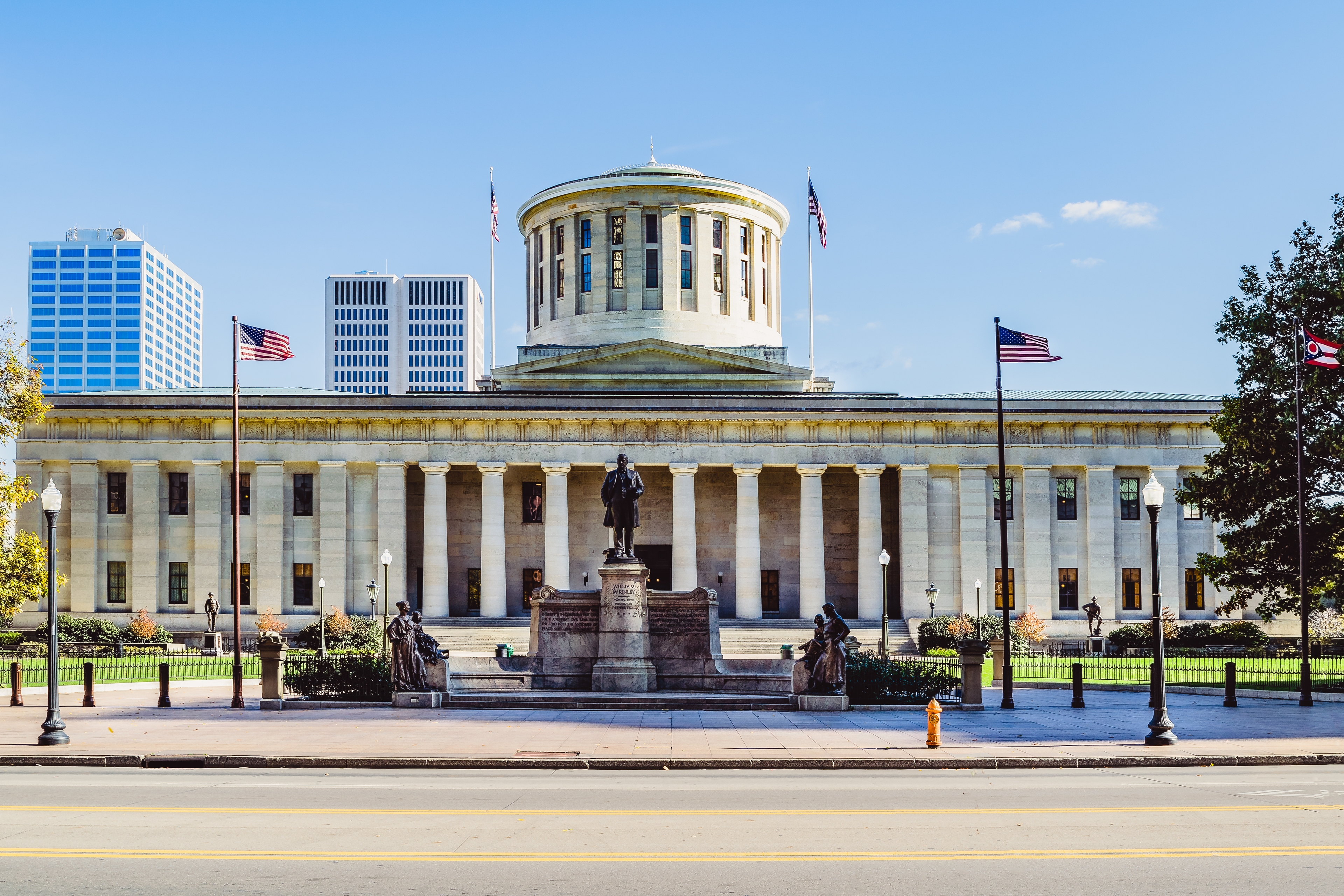 Front view of the Ohio Statehouse in Columbus, featuring its iconic rotunda, neoclassical columns, and surrounding statues under a clear blue sky.