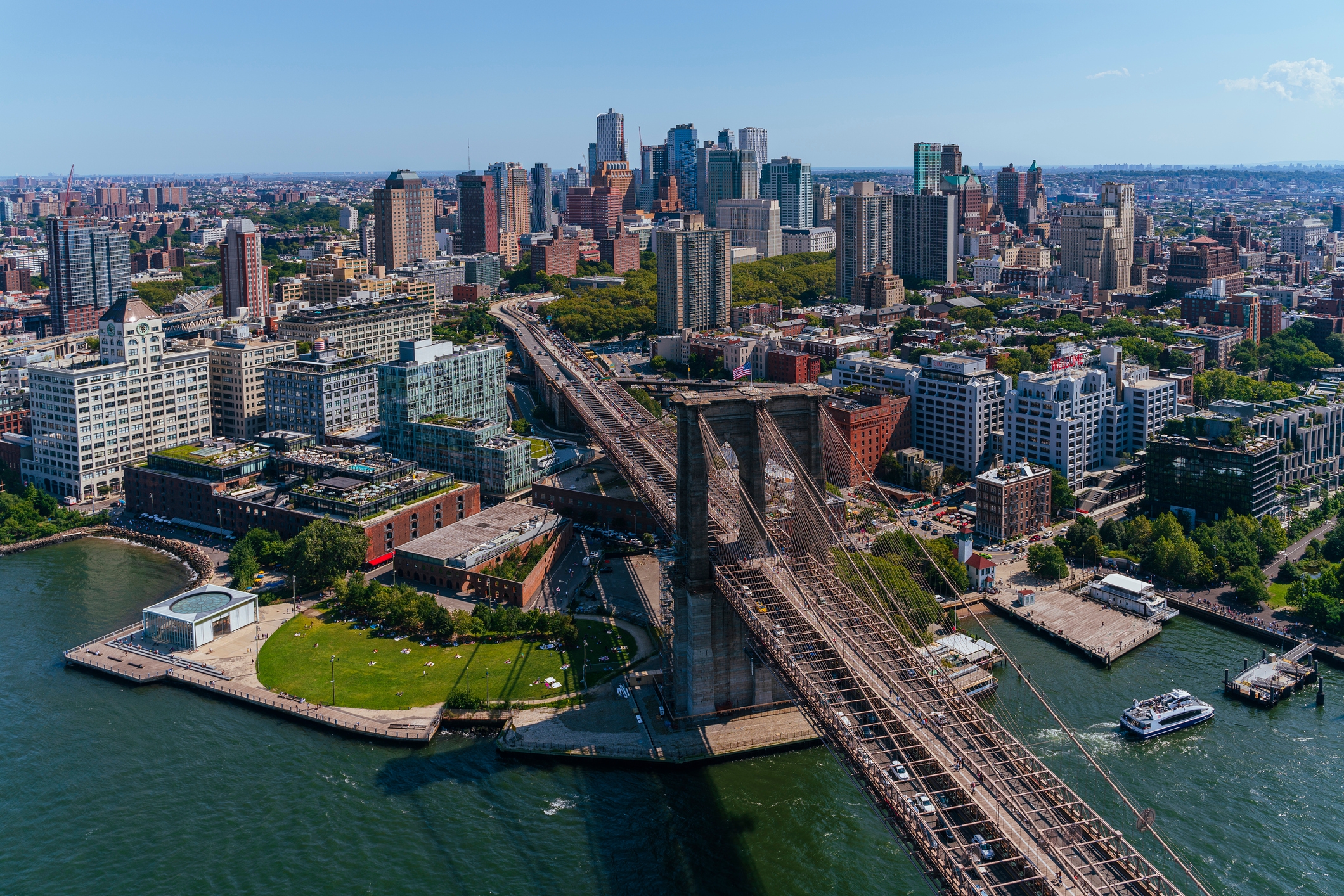 An aerial view of Brooklyn, New York, highlighting the Brooklyn Bridge extending over the East River towards downtown Brooklyn. The scene features parks and waterfront areas in the foreground, with a backdrop of high-rise office buildings, residential complexes, and tree-lined streets under a clear blue sky.