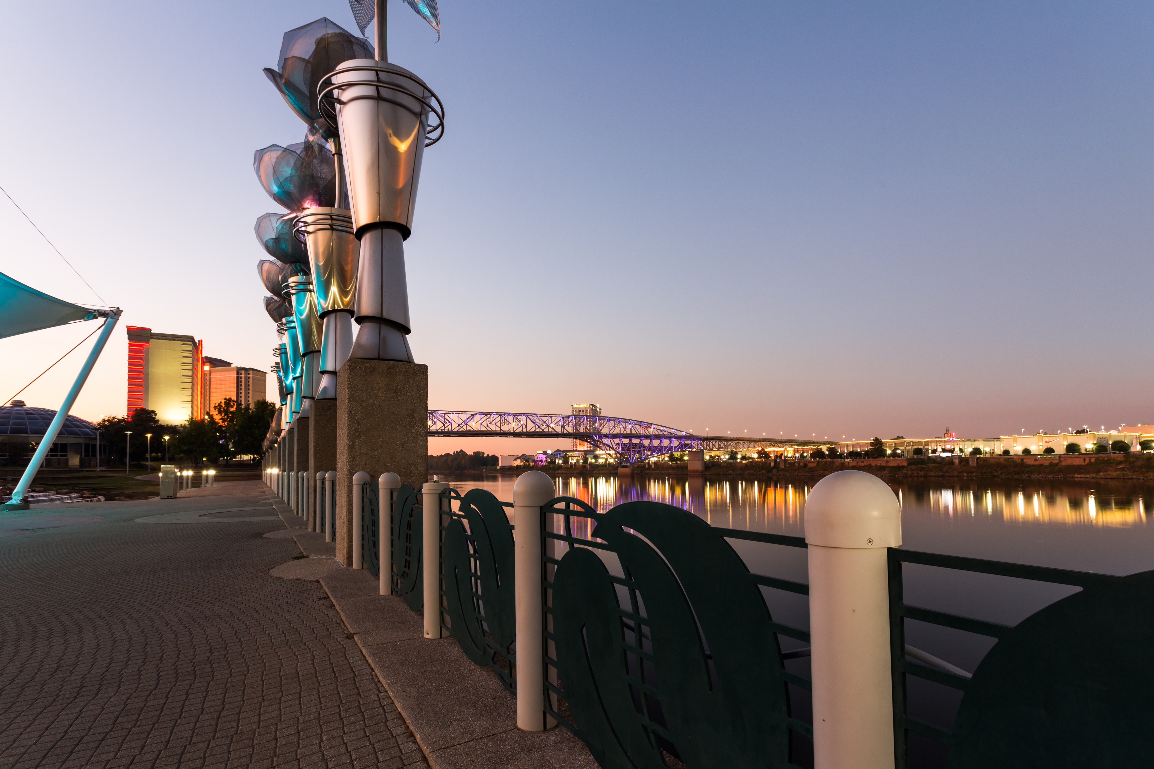 Evening view of the Shreveport riverfront, with illuminated sculptures and a glowing bridge reflecting in the Red River under a twilight sky.