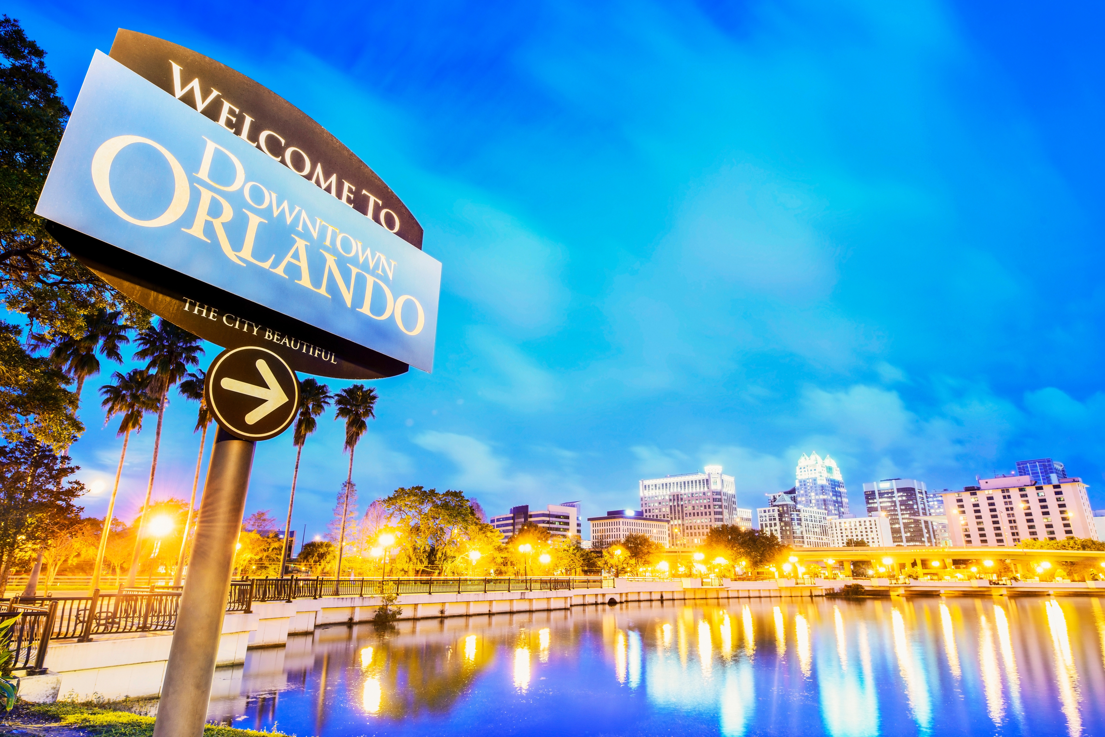 Welcome sign for Downtown Orlando at dusk, with city lights reflecting on the water and a vibrant skyline in the background.