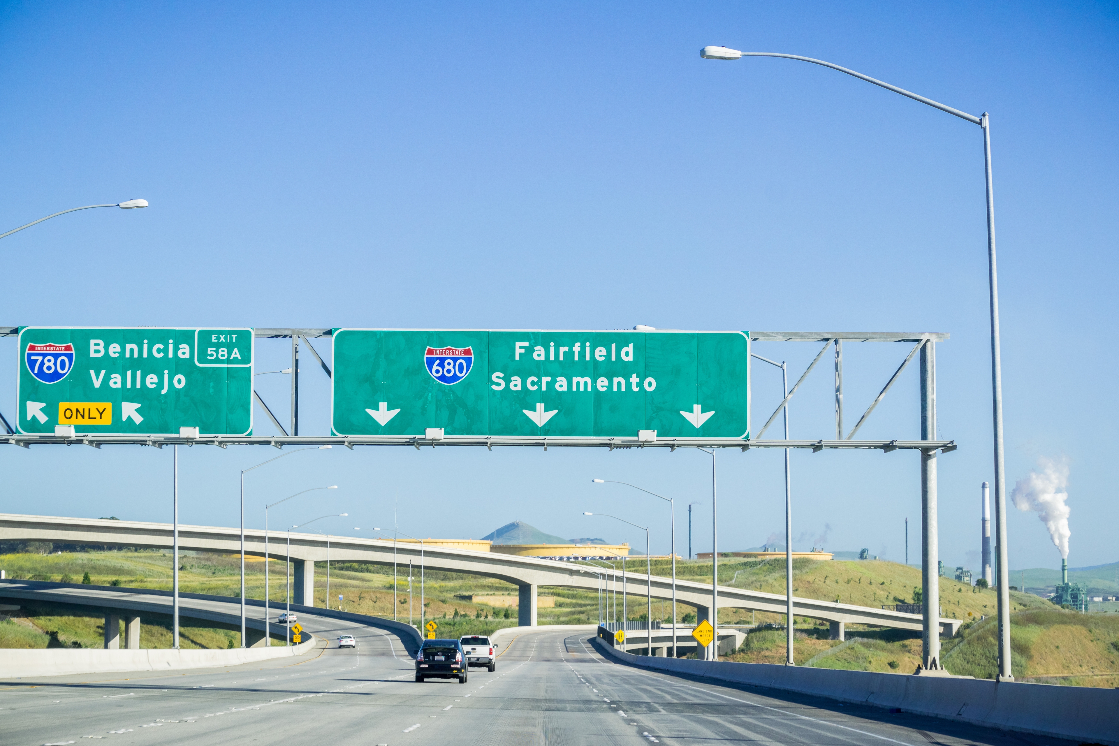 A highway scene near Fairfield, California, showing directional signs for Interstates 680 and 780 leading to destinations like Benicia, Vallejo, and Sacramento, with an overpass and industrial smokestacks in the distance under a clear blue sky.