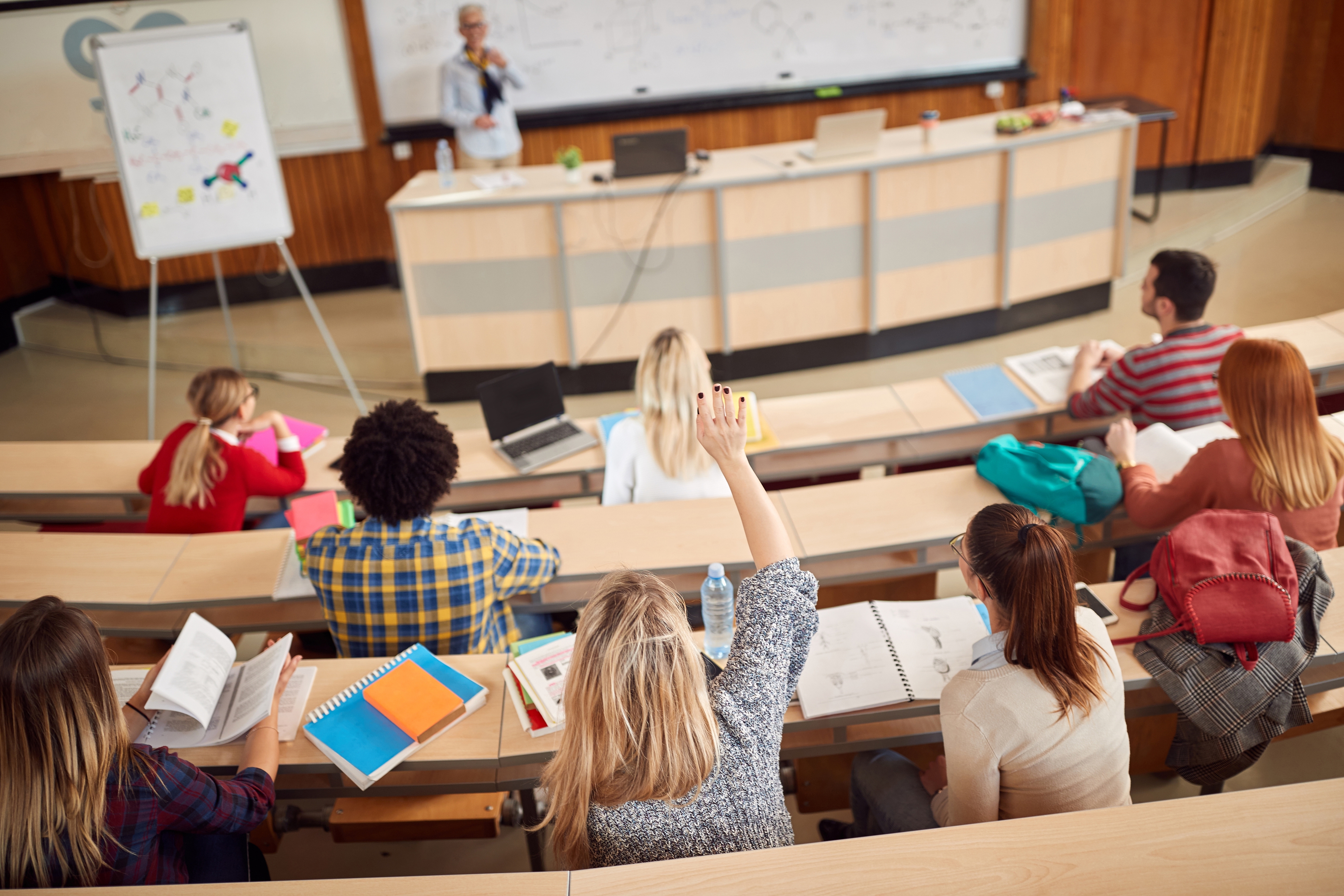 A diverse group of university students attending a lecture in a modern classroom. A student in the foreground raises their hand to ask a question, while others take notes or reference textbooks. The instructor stands at the front near a whiteboard and lectern, engaging with the class.