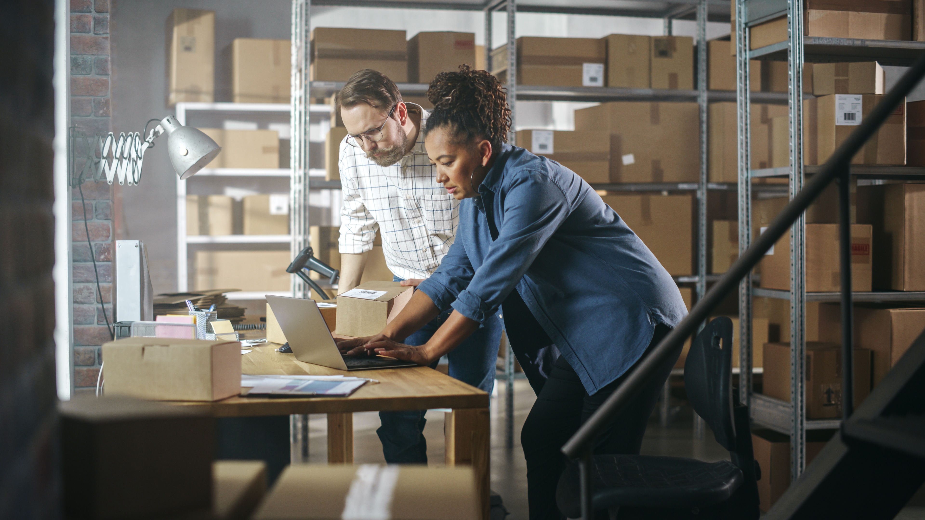 Two workers in a warehouse setting, surrounded by shelves filled with cardboard boxes. One person is using a laptop while the other holds a package, indicating they are likely managing inventory or processing orders.