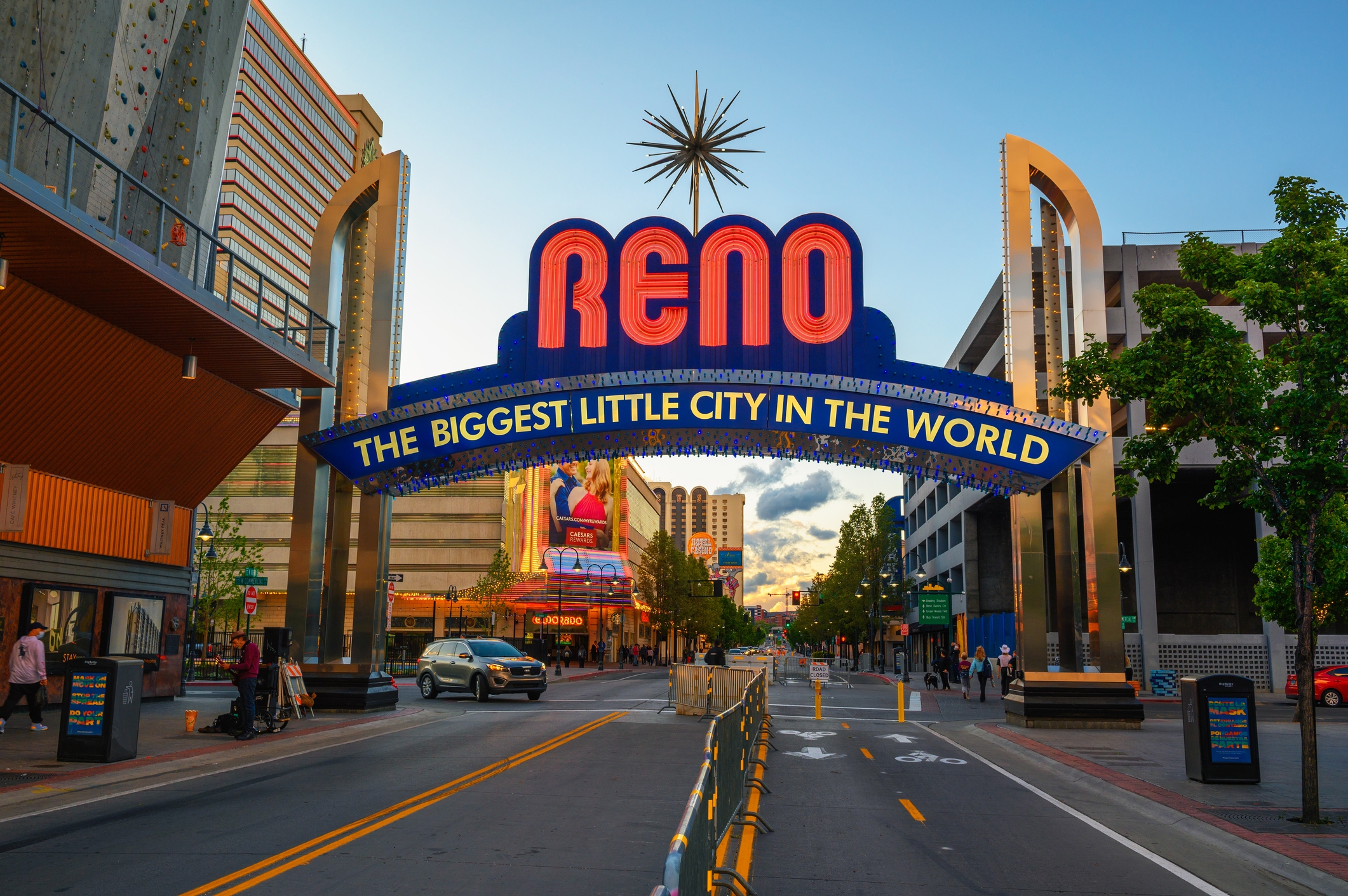 The Reno Arch in downtown Reno, Nevada, with bold red lettering reading 'Reno' and the slogan 'The Biggest Little City in the World' illuminated in lights. The street below features cars and pedestrians, surrounded by buildings under a partly cloudy evening sky.