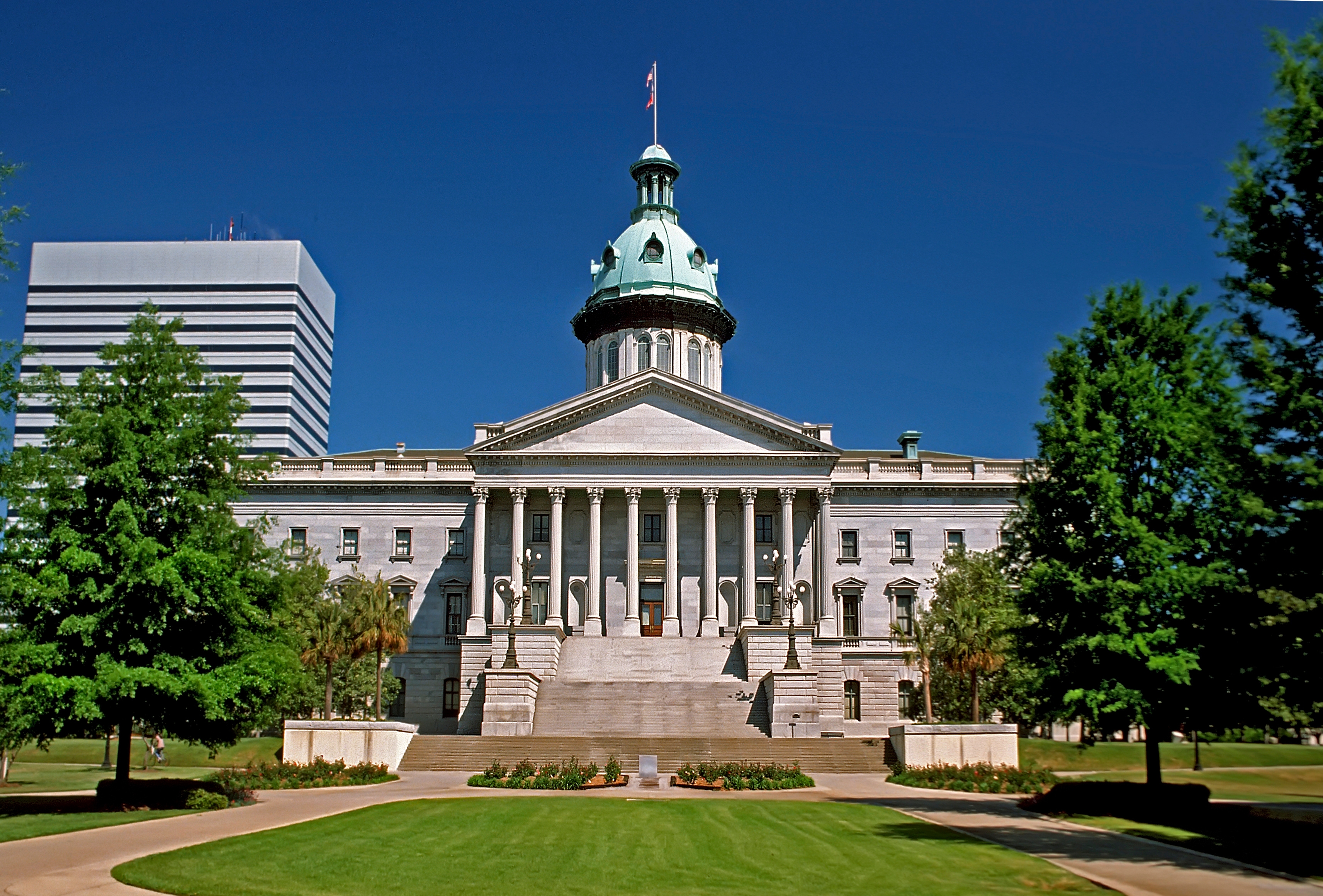 Front view of the South Carolina State House in Columbia, featuring a grand staircase, Corinthian columns, and a distinctive copper dome under a deep blue sky.
