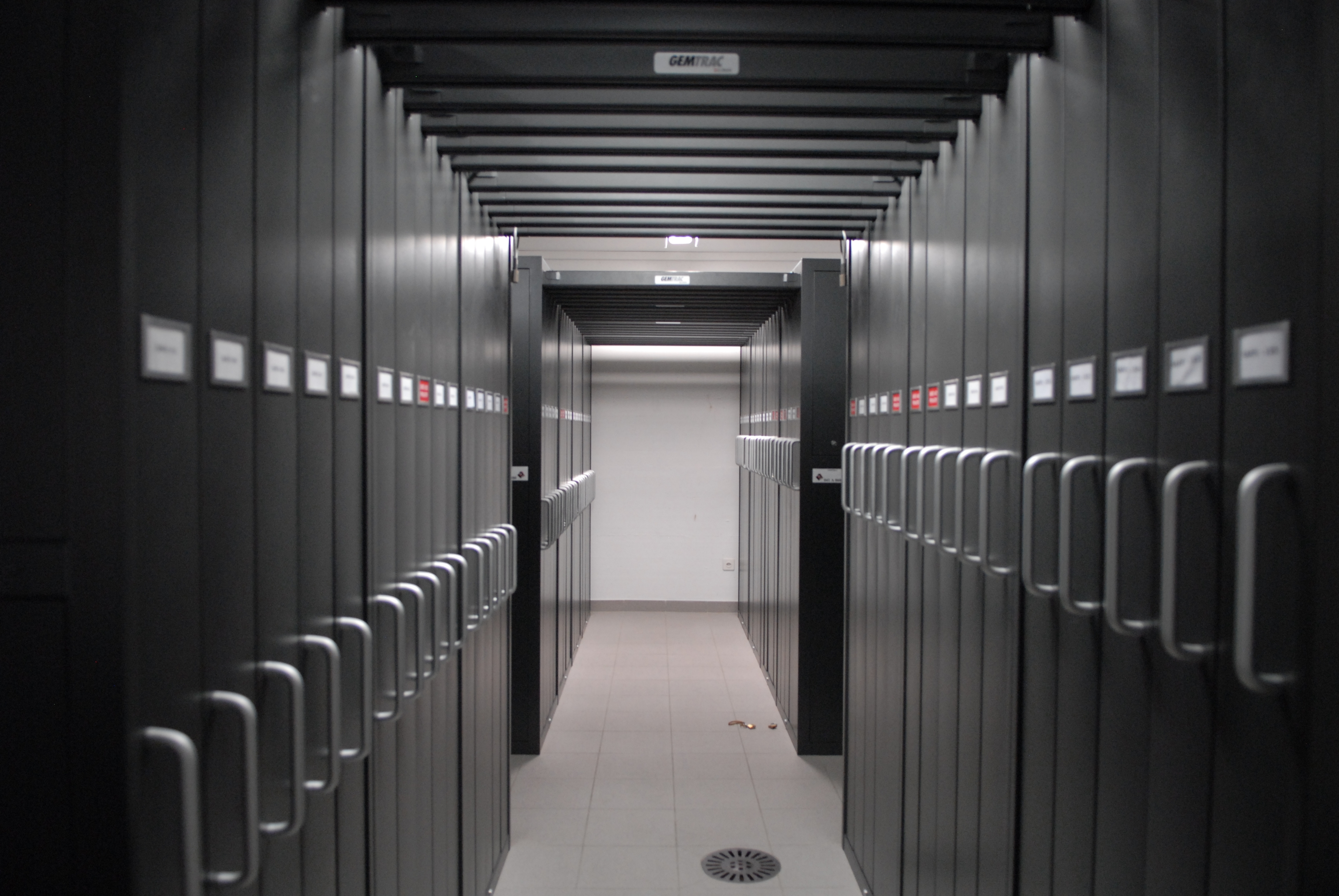 A narrow aisle between large, high-density storage shelves in a modern archival facility. The shelves are labeled and equipped with handles, designed for efficient storage and retrieval of documents or files.