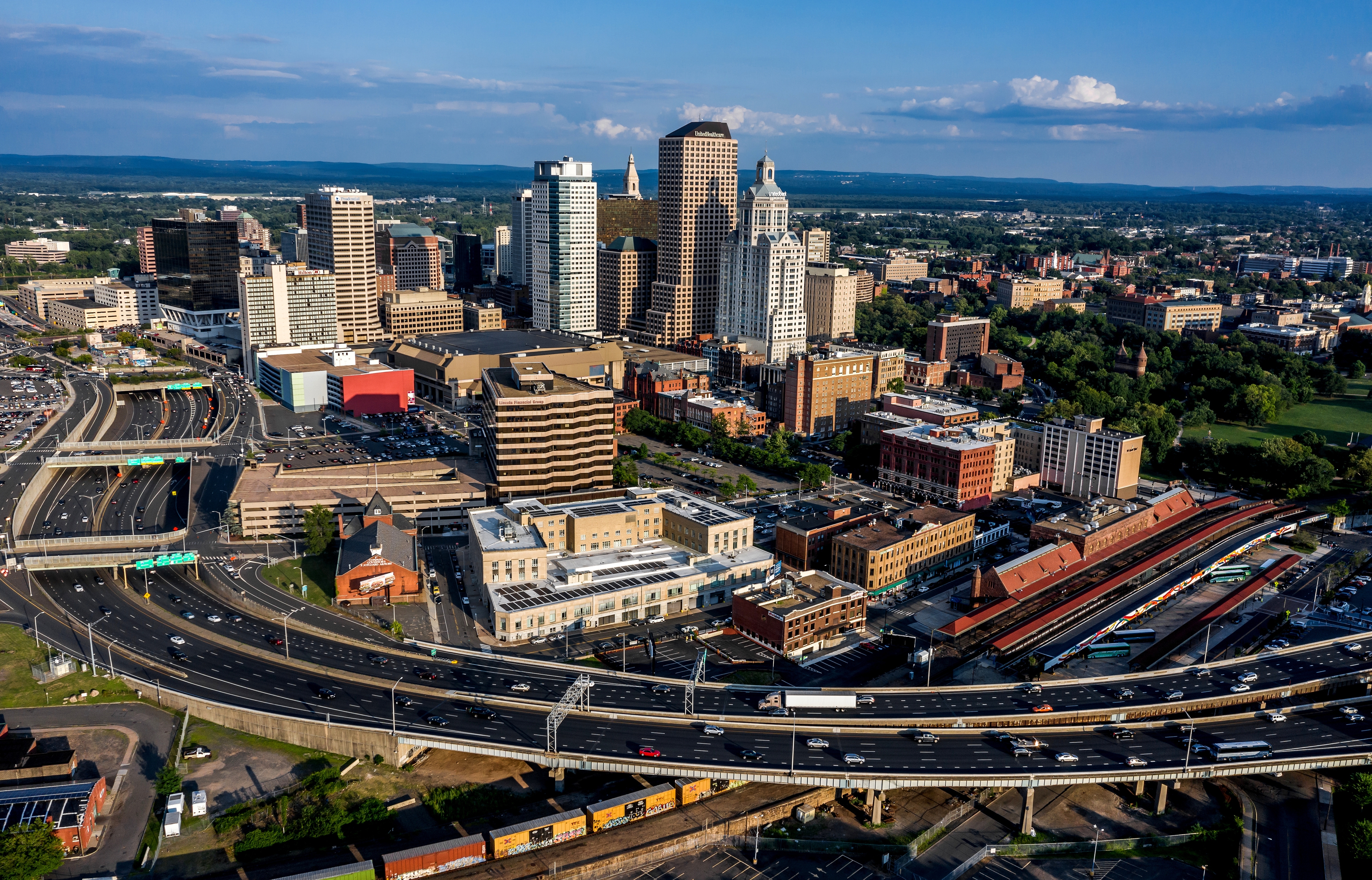 An aerial view of downtown Hartford, Connecticut, showcasing a mix of modern and historic architecture, intersecting highways, and a train station, with lush greenery and a clear blue sky in the background.