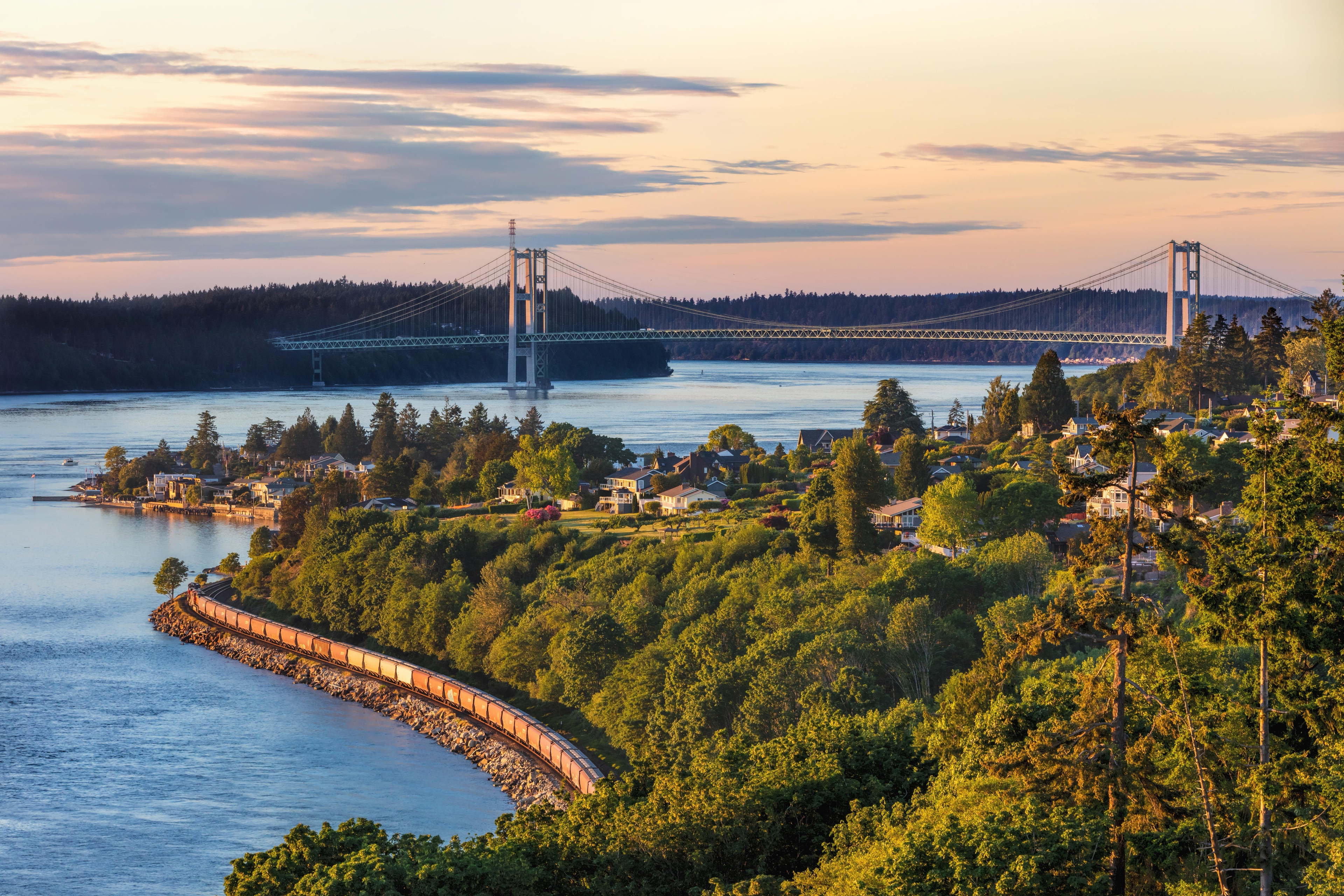 Scenic view of the Tacoma Narrows Bridge in Washington at sunset, with a freight train curving along the shoreline below and a neighborhood nestled among lush greenery in the foreground.