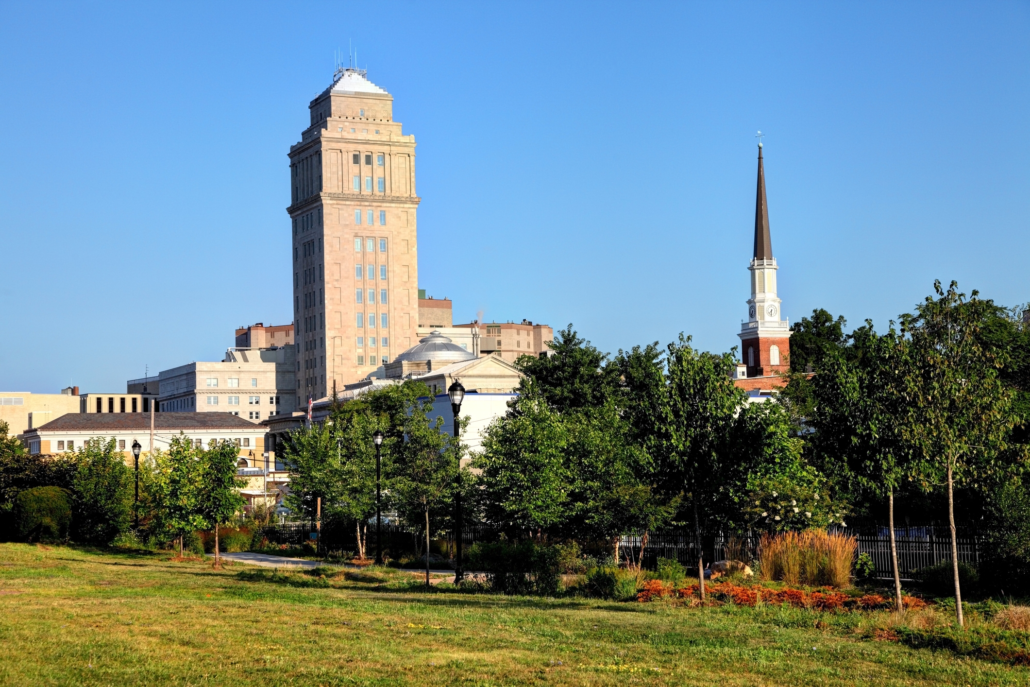 A view of Elizabeth, New Jersey, featuring a tall beige office building and a church with a pointed steeple rising above trees and a grassy park in the foreground. The scene is set under a bright blue sky.