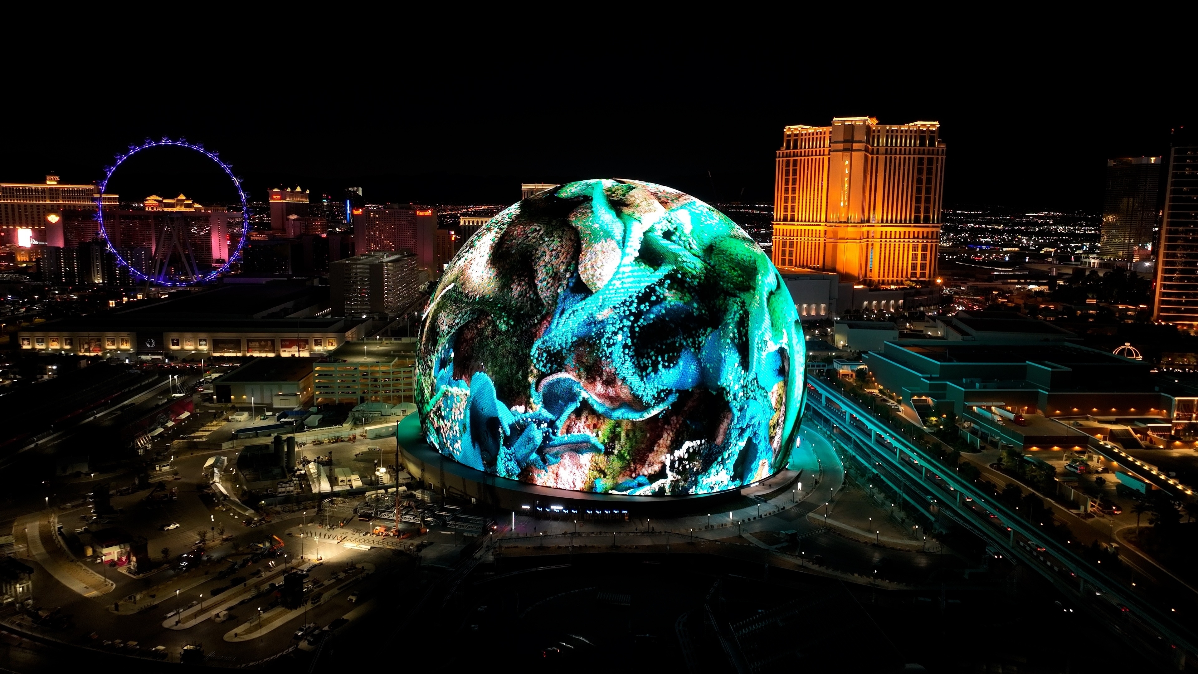 Night view of the Las Vegas Sphere illuminated with digital visuals, surrounded by city lights and iconic landmarks like the High Roller and Palazzo.