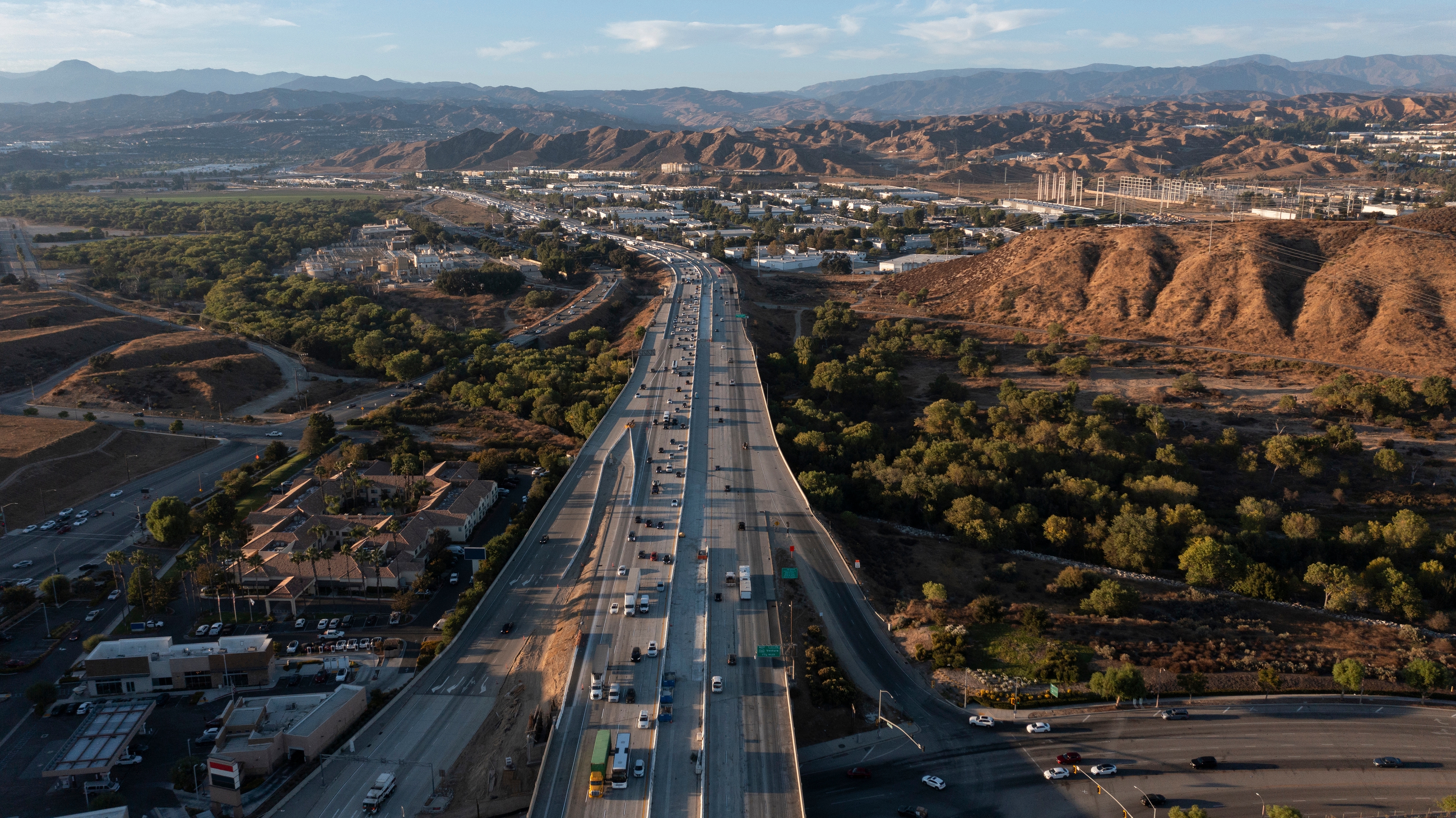 Aerial view of a busy freeway cutting through Santa Clarita, California, with surrounding commercial areas, greenery, and rugged hills in the background.