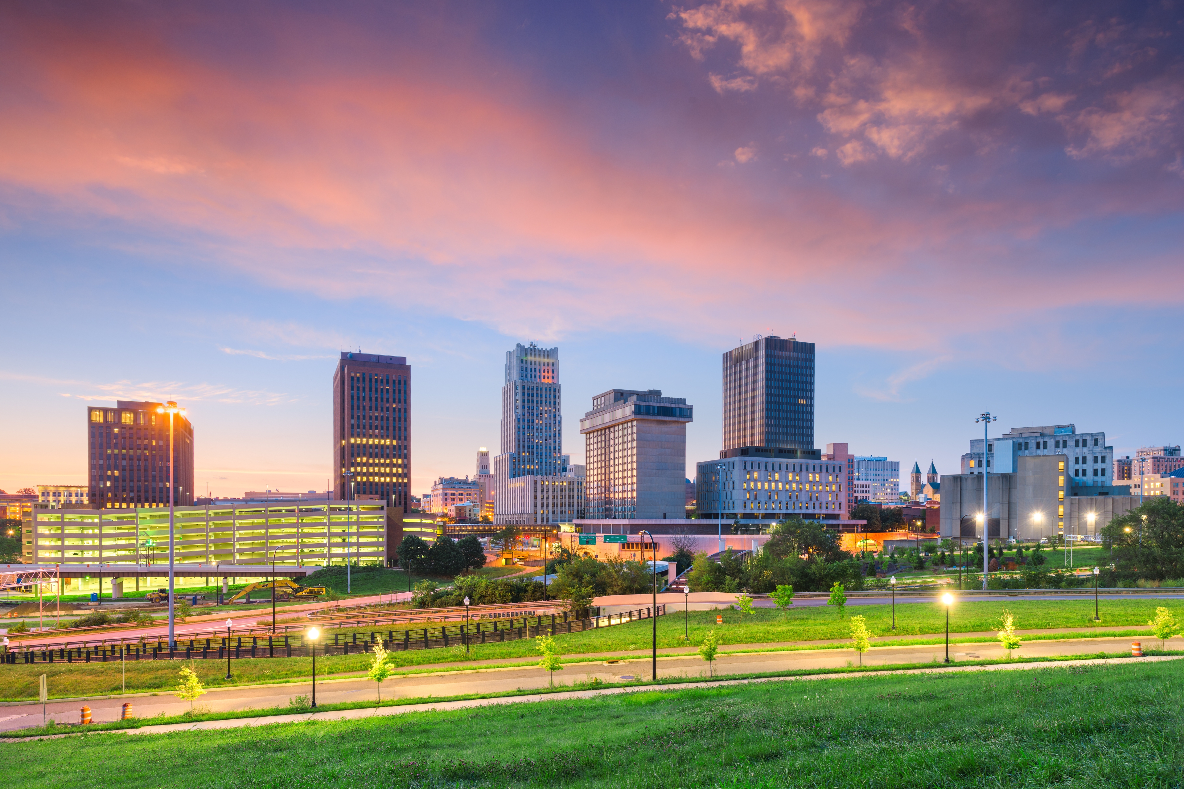 Akron, Ohio skyline at dusk, with a dramatic pink and orange sky illuminating the city. Modern and historic buildings create a dynamic urban scene, with green grass and pathways in the foreground.