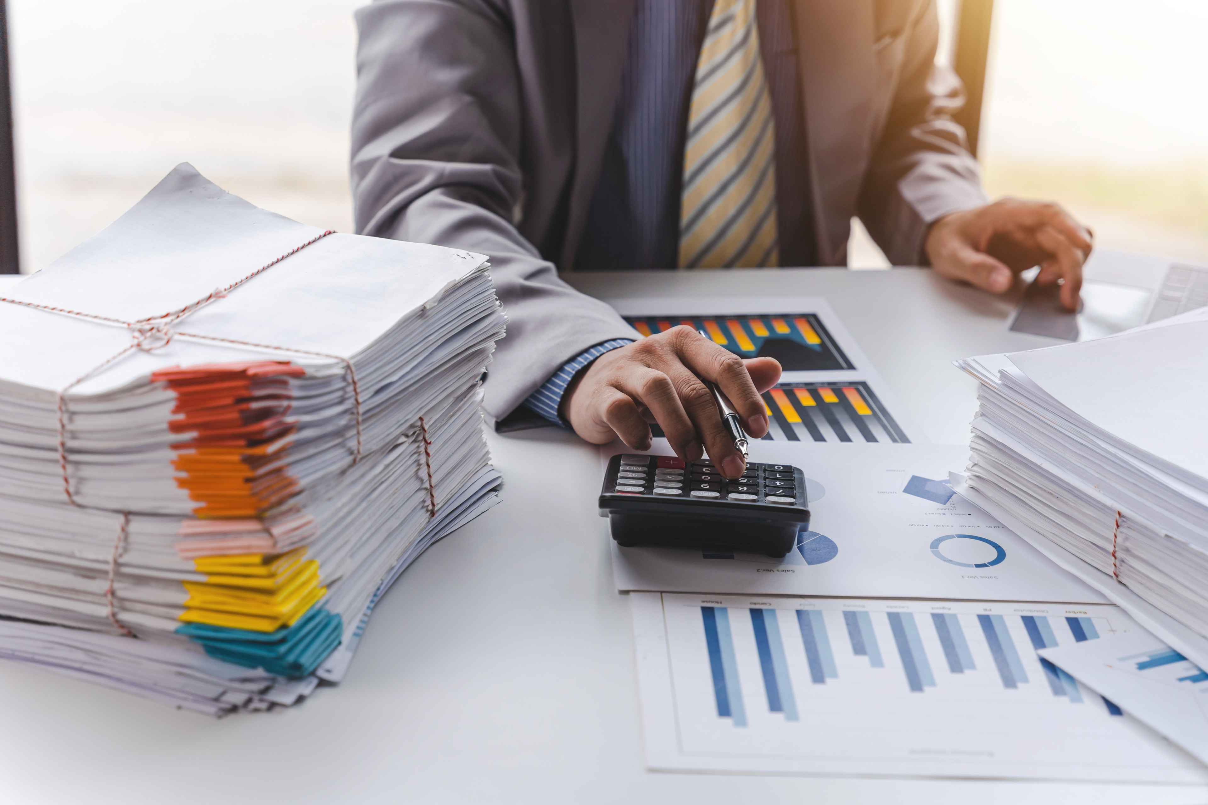 A person in business attire using a calculator at a desk, surrounded by stacks of documents and financial charts. The documents are tied with strings, and the charts display bar and pie graphs, indicating financial analysis or auditing work.