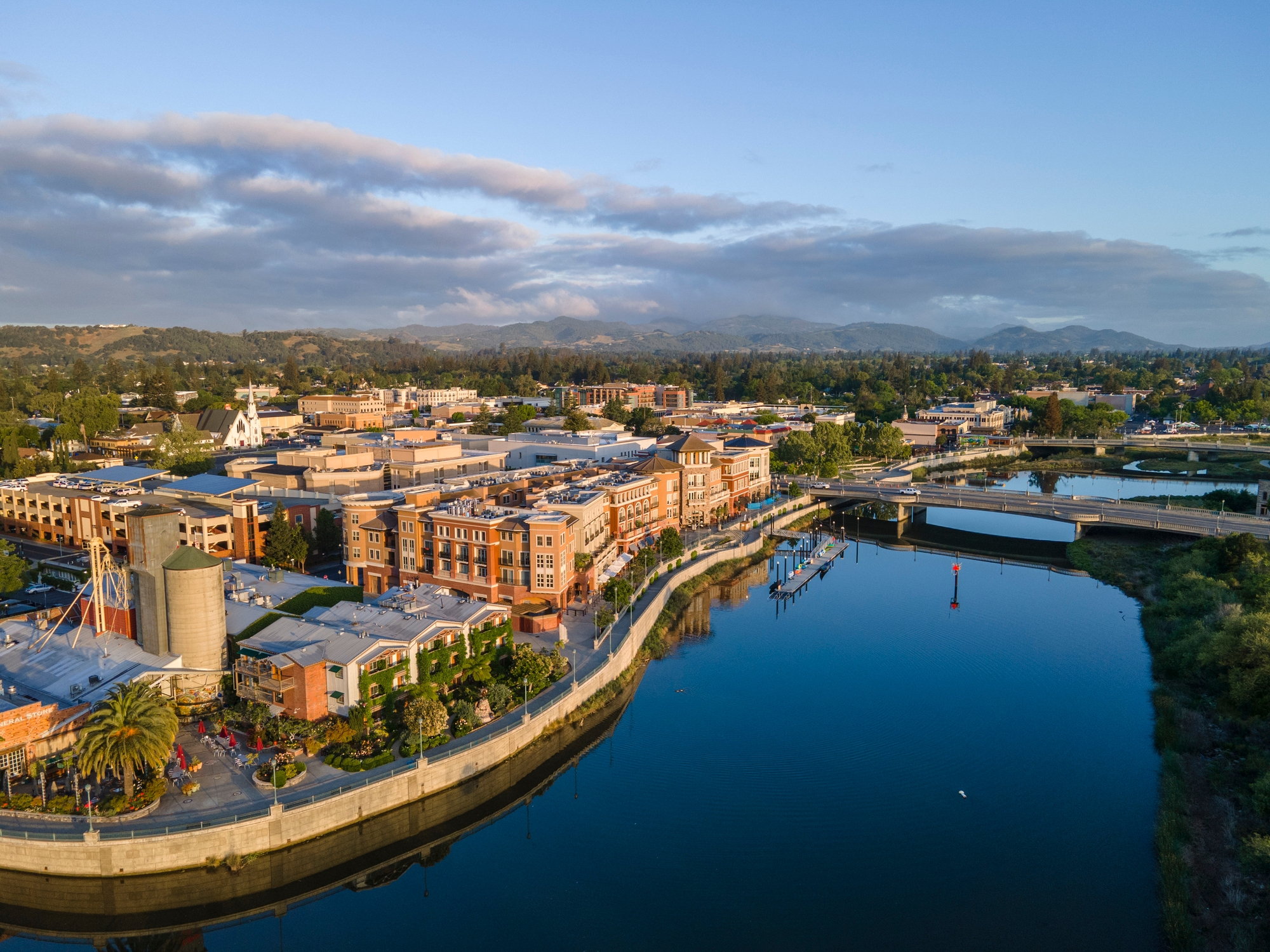 An aerial view of Napa, California, showcasing its downtown area with buildings along the riverfront. A calm river reflects the structures, bridges, and surrounding lush green hills under a partly cloudy sky.