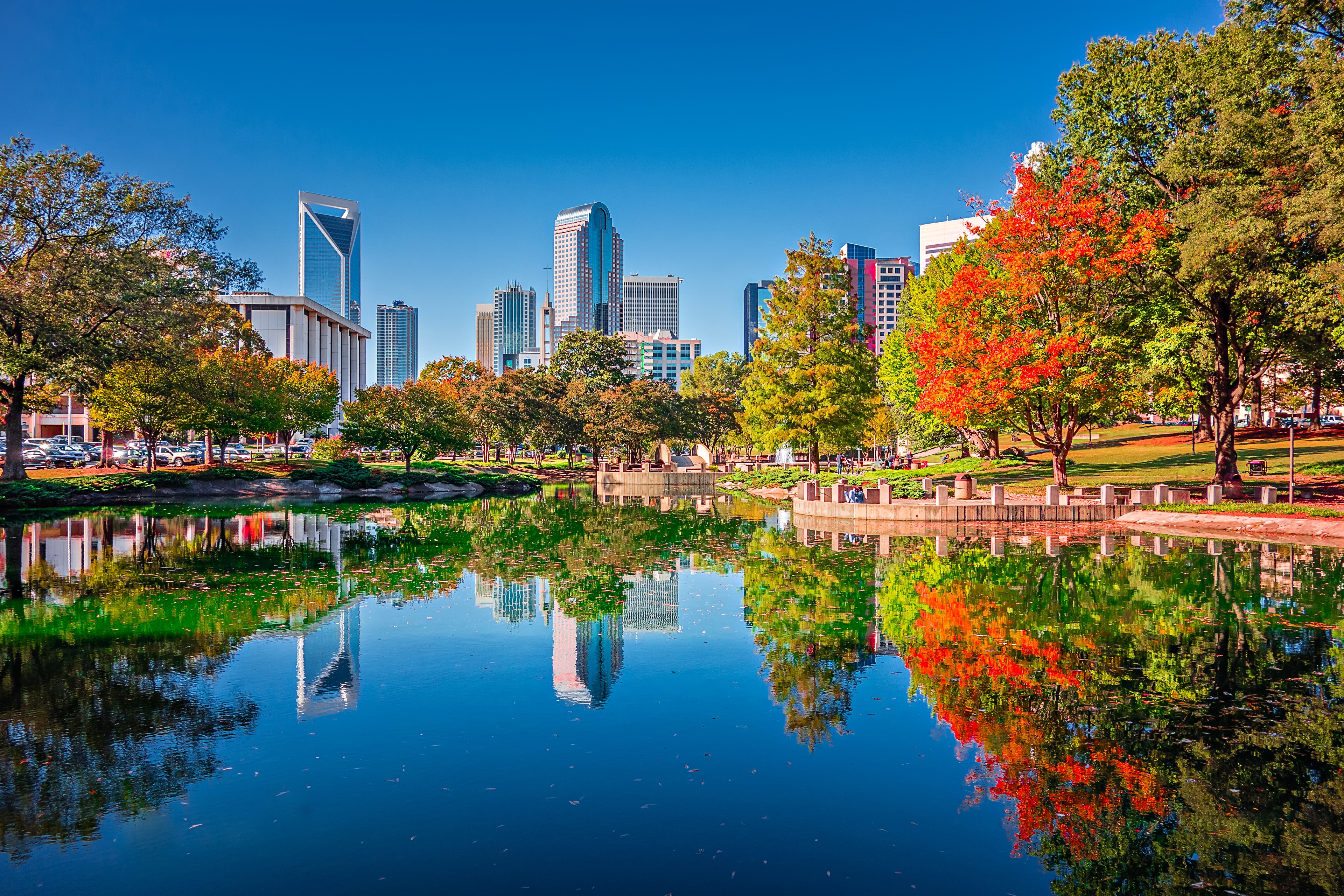 Charlotte, North Carolina skyline view from Marshall Park, featuring autumn-colored trees and skyline reflections in the pond under a clear blue sky.