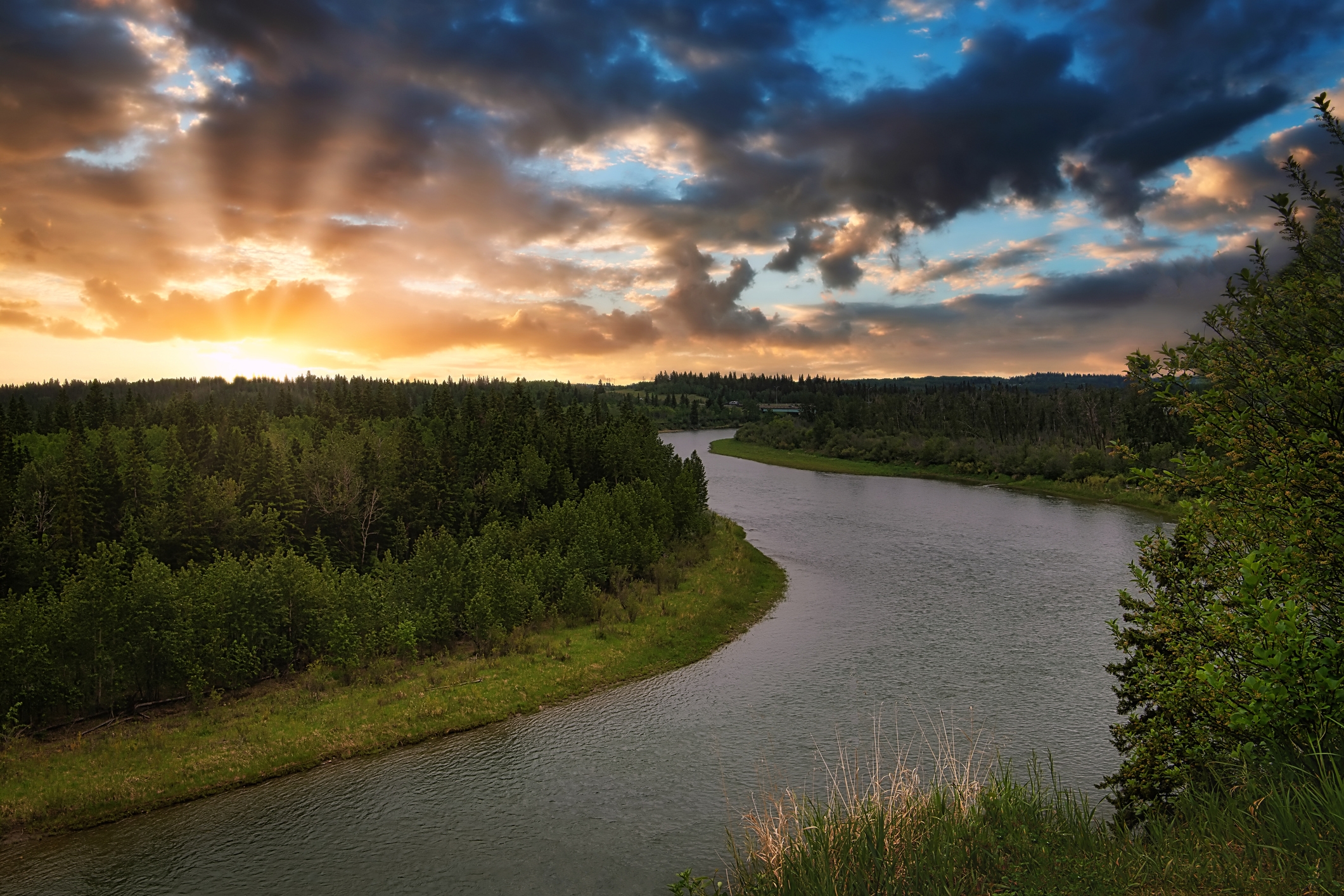 A sunset over the Red Deer River in Alberta, Canada, with golden rays piercing through clouds above a lush forested landscape and the winding river.