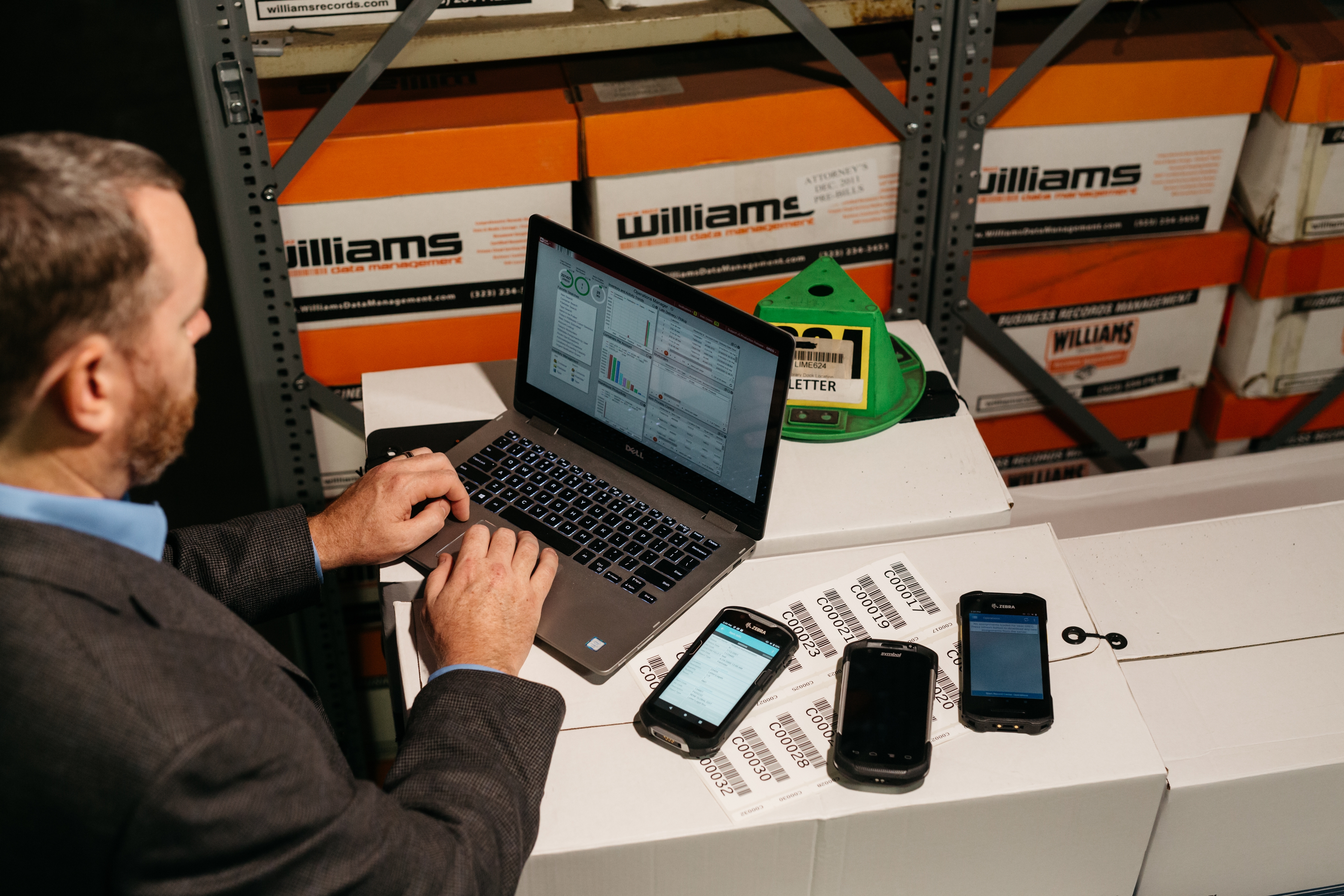 A man working on a laptop in a warehouse environment, reviewing records management software. Multiple handheld barcode scanners are placed next to the laptop, and labeled boxes are stacked on shelves in the background, indicating a focus on inventory or records management.