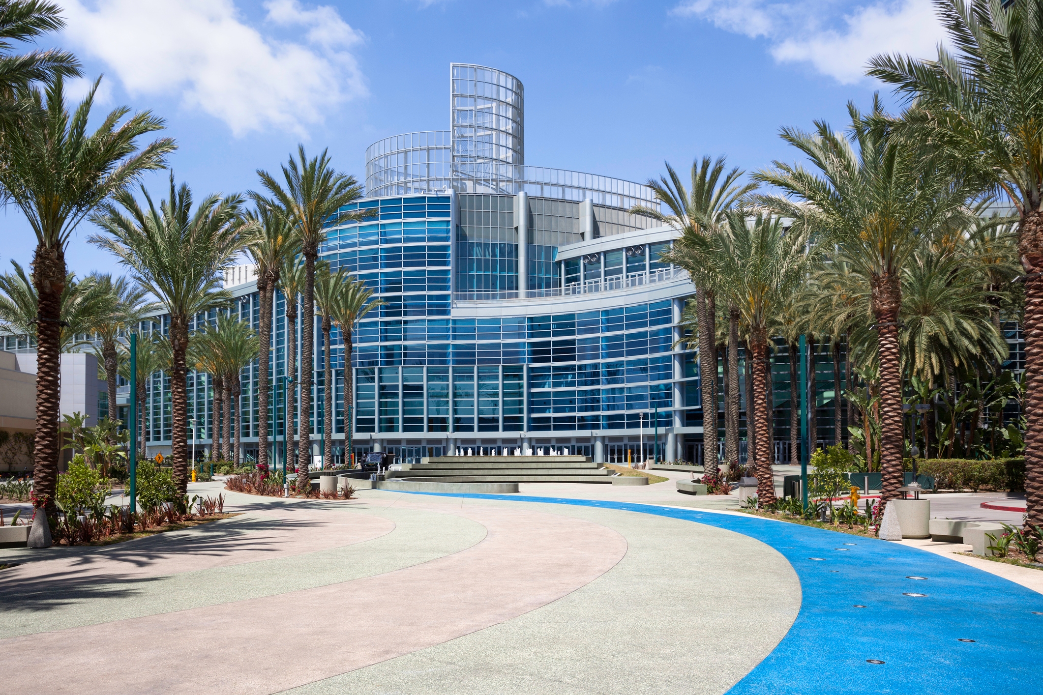 Modern glass façade of the Anaheim Convention Center in California, framed by palm trees and a vibrant pedestrian path under a clear blue sky.