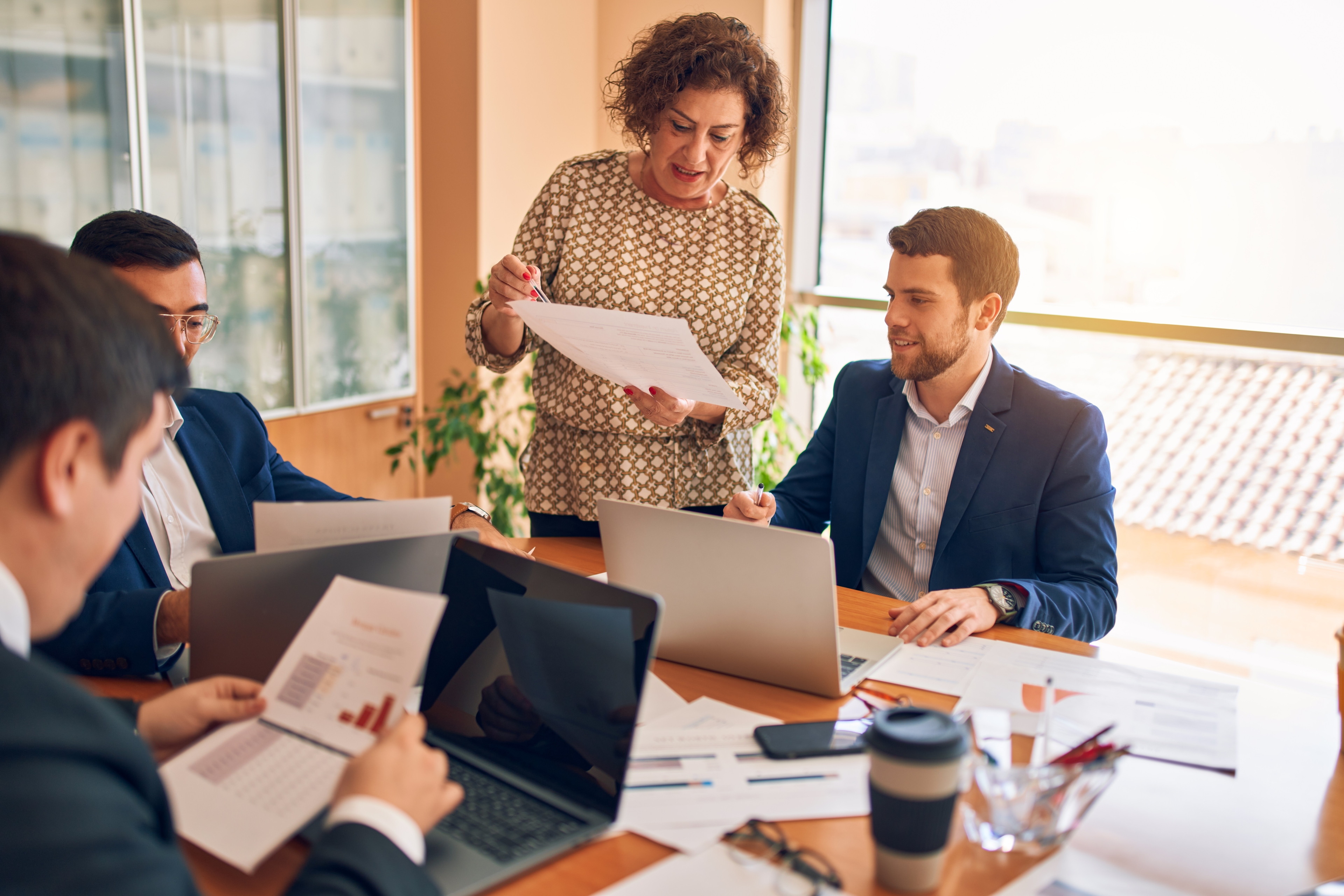 Business meeting in a bright office with five professionals discussing documents and working on laptops around a table.