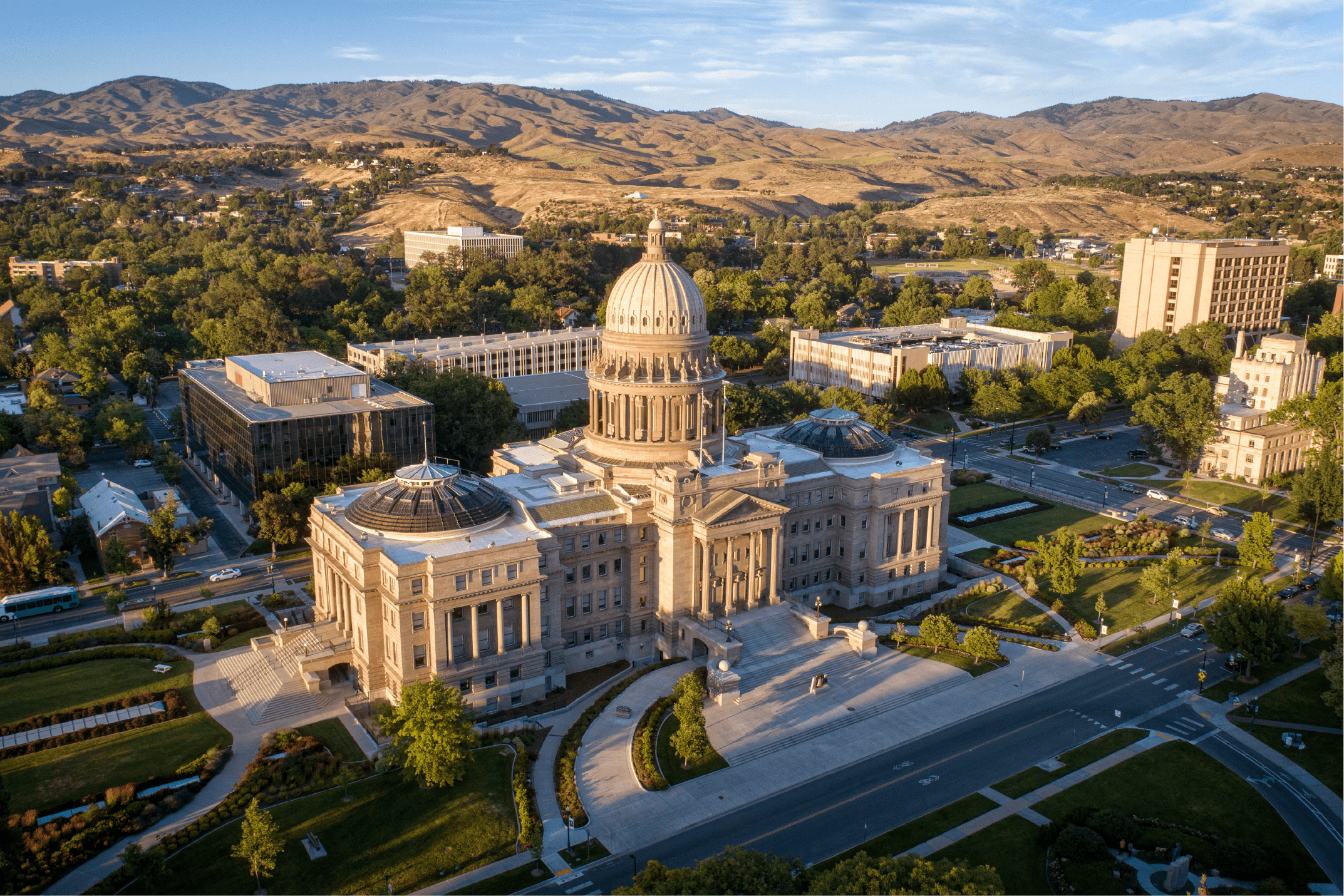 An aerial view of the Idaho State Capitol building in Boise, surrounded by trees, city structures, and rolling hills under a clear blue sky.