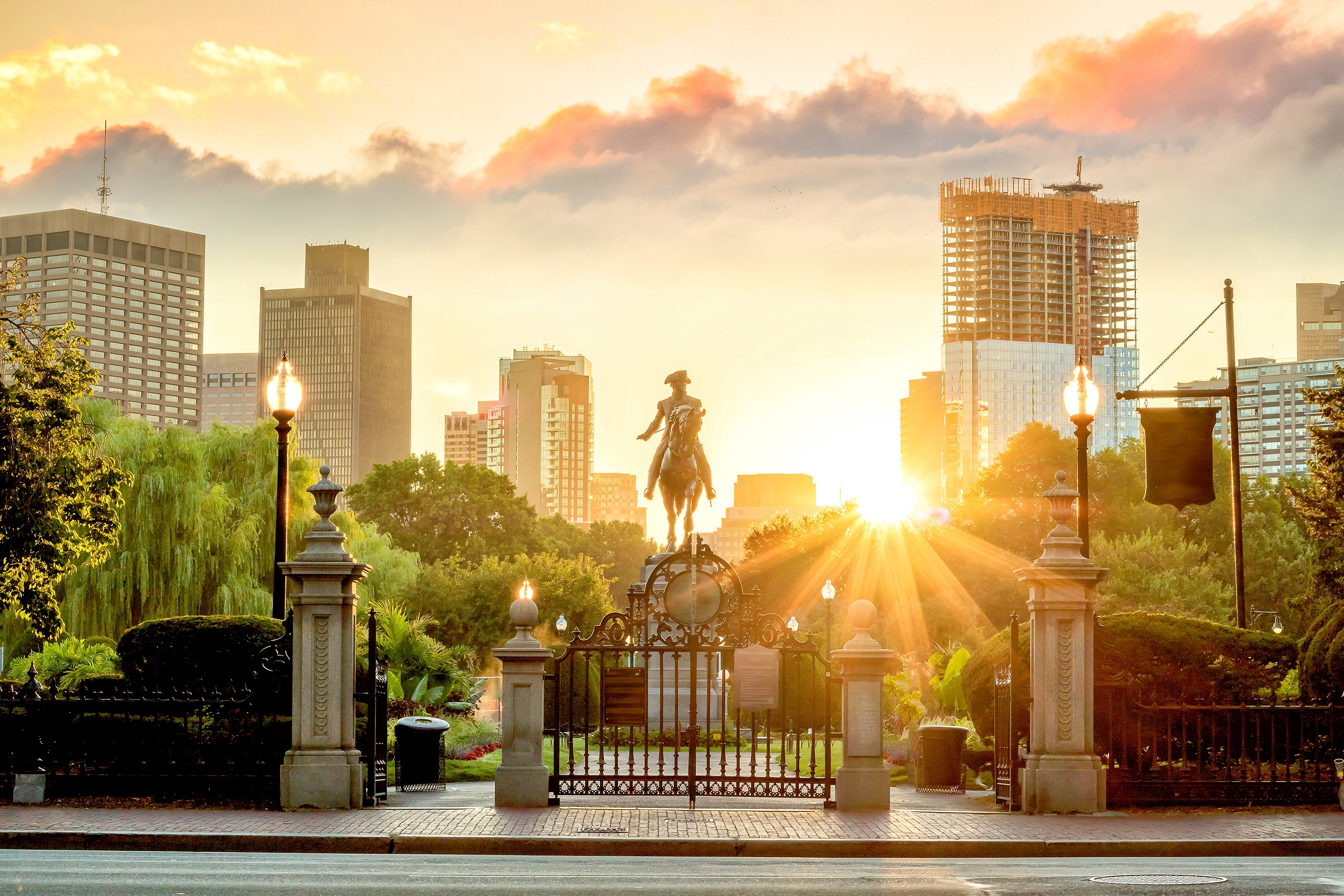 Sunrise over Boston Public Garden with a silhouette of the George Washington statue, framed by historic gates and modern skyline architecture.