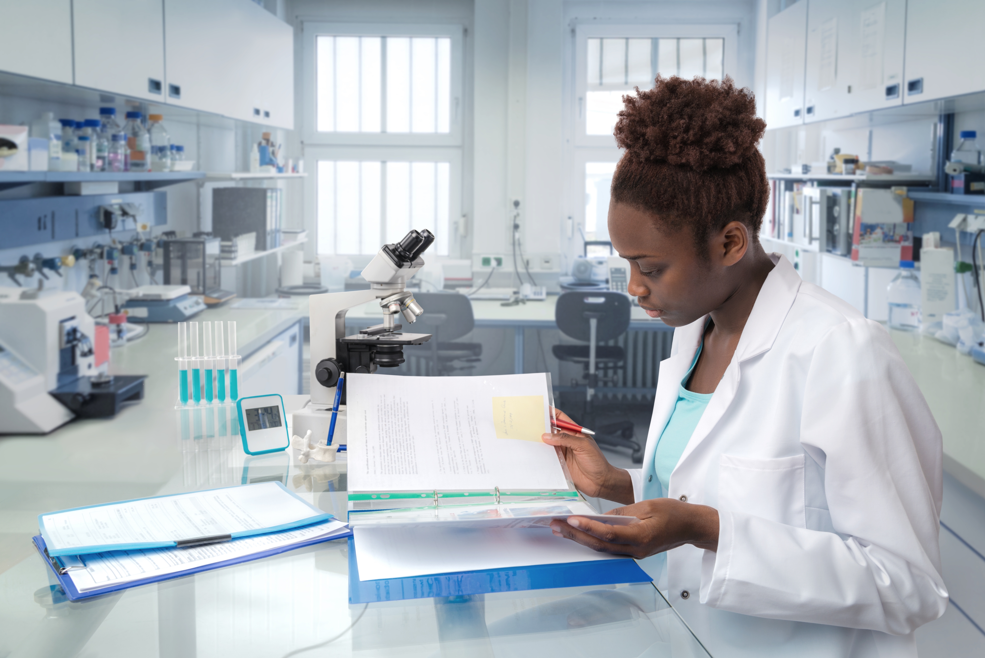 Female scientist in a lab coat reviews documents at a desk with a microscope and test tubes in a well-equipped laboratory
