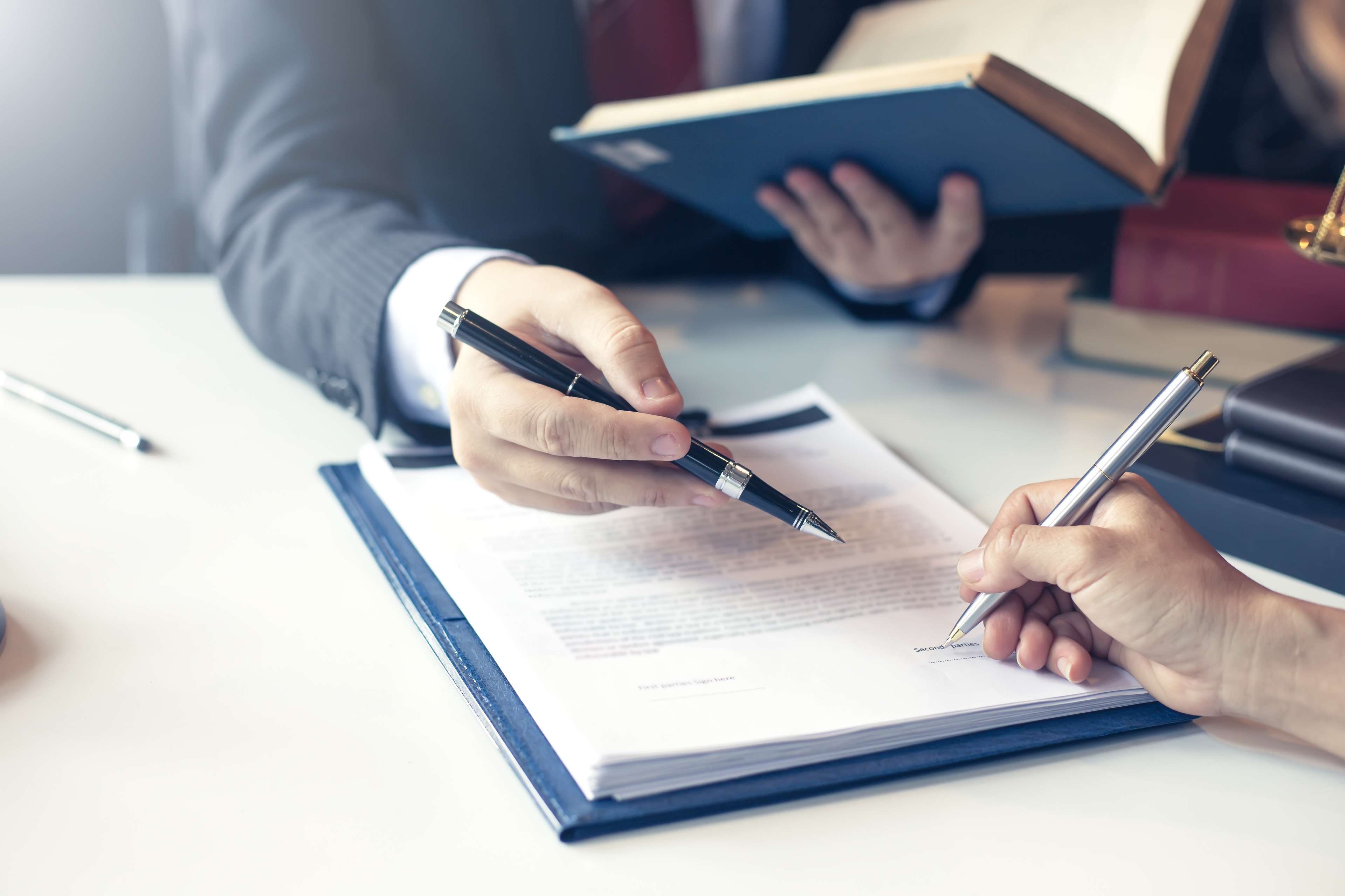 Two professionals in business attire working on a legal or official document. One person is holding a book, while the other points to a section on the document with a pen, guiding the other to sign. The scene suggests a formal agreement or contract review process.