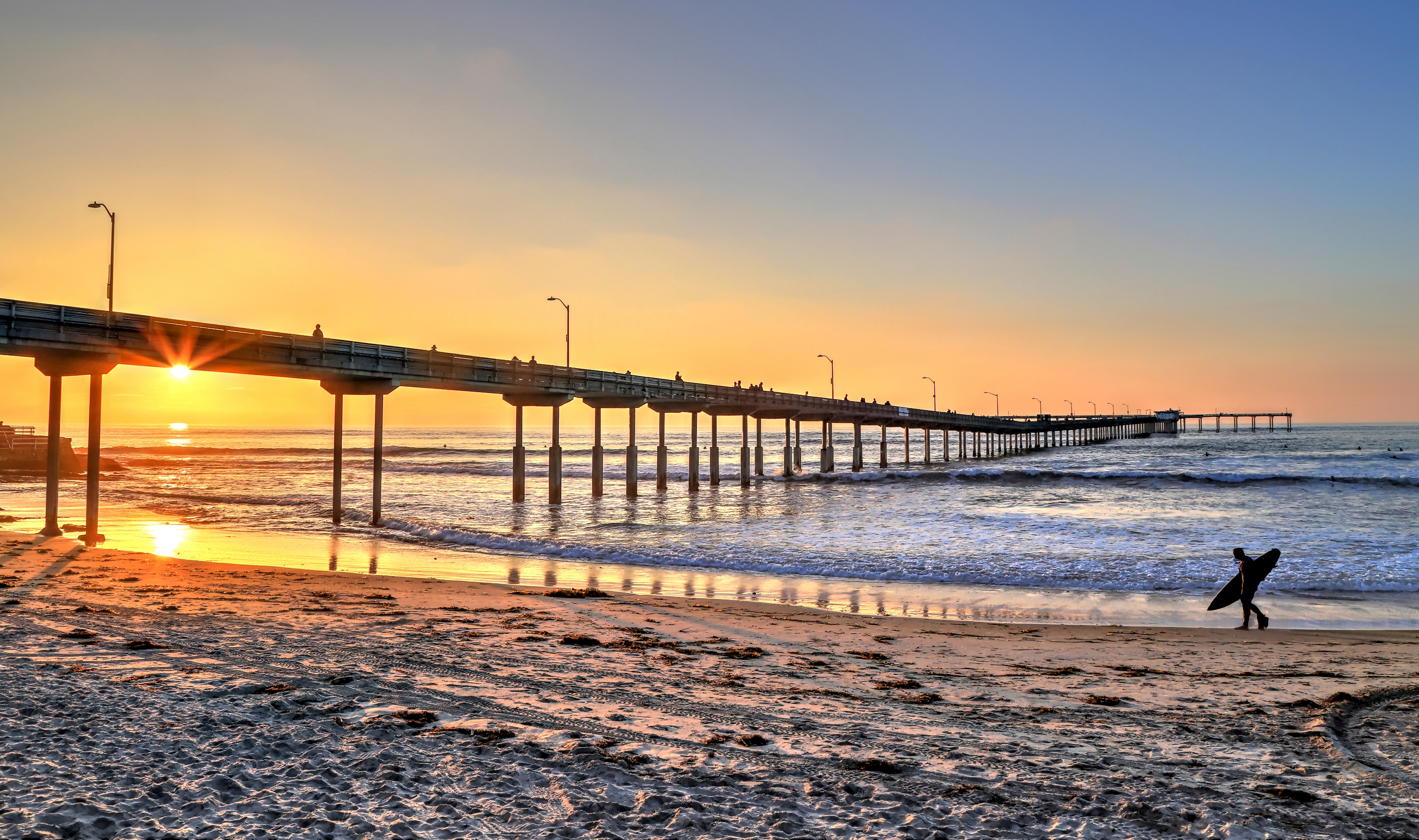 Ocean Beach Pier at sunset with a silhouetted surfer walking along the shoreline, capturing San Diego's laid-back coastal vibe.