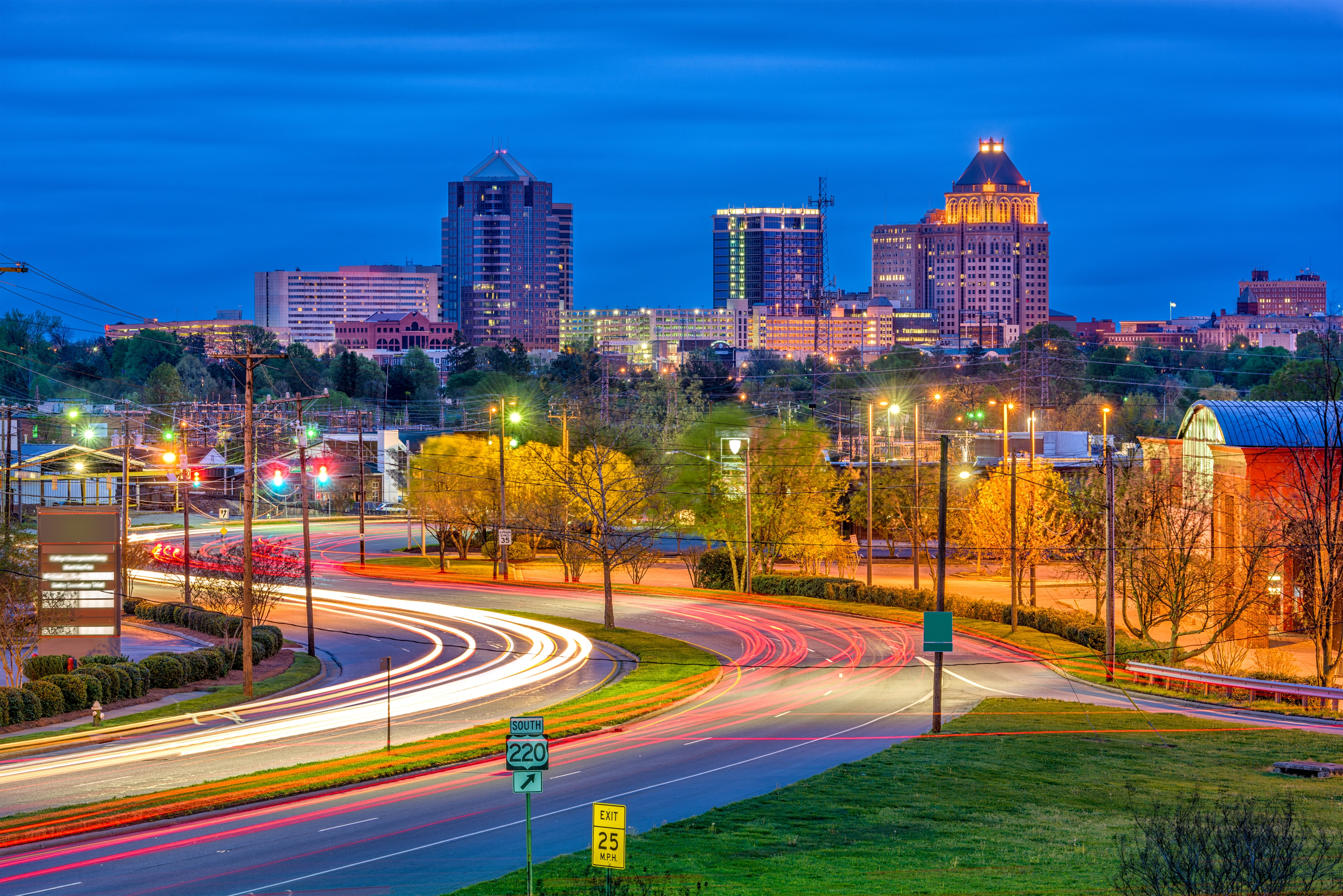 Nighttime cityscape of Greensboro, North Carolina, featuring light trails from Highway 220 traffic and the brightly lit skyline with iconic buildings.

