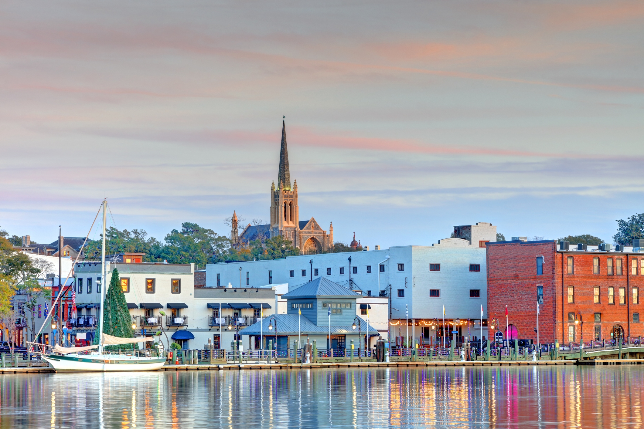 Sunset over Wilmington, North Carolina’s riverfront, featuring historic architecture, a sailboat, and the iconic spire of St. James Episcopal Church reflecting on the water.