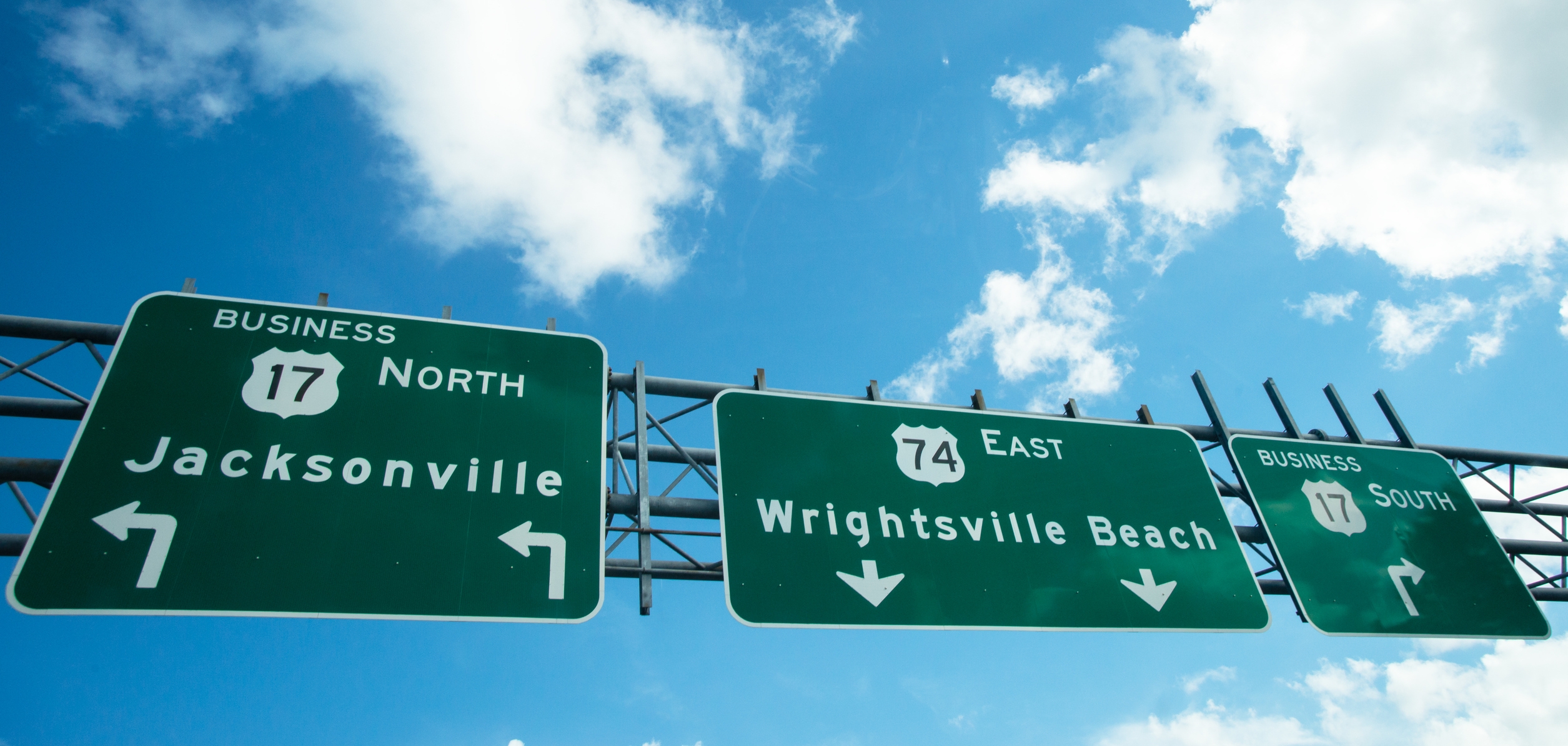 Overhead road signs on U.S. Route 17 and U.S. Route 74 guide drivers toward Jacksonville, North Carolina, and Wrightsville Beach against a bright blue sky.