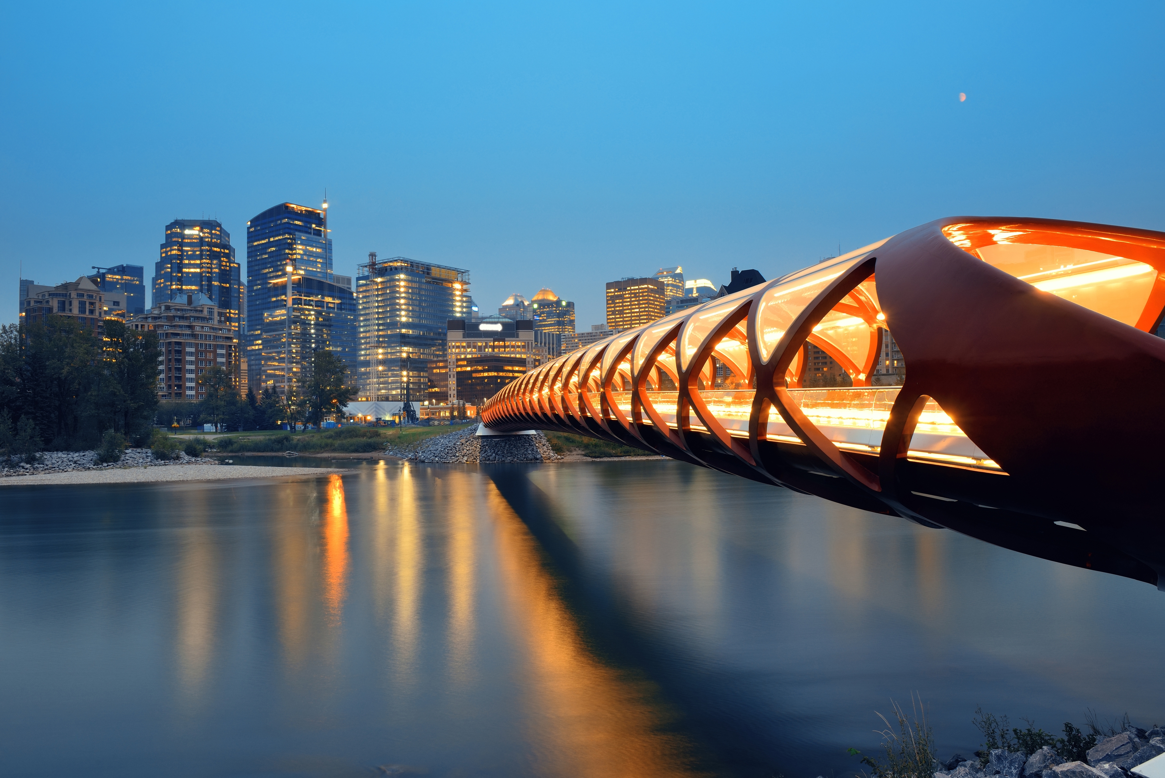 The illuminated Peace Bridge in Calgary spans the calm Bow River at dusk, with the city's skyline glowing in the background under a deep blue sky.