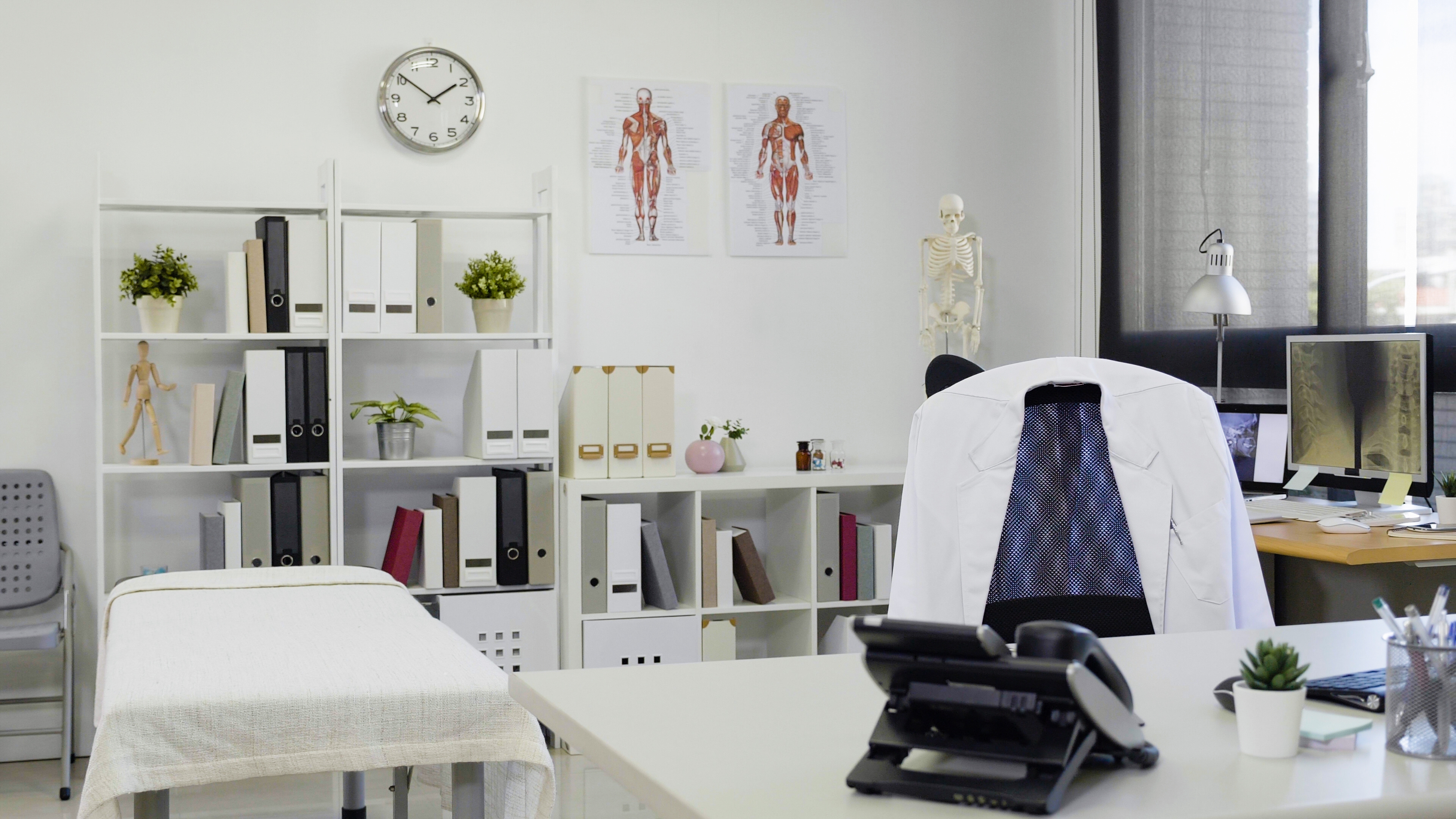 Doctor's office with a desk, medical posters, anatomical model, bookshelves, and examination table