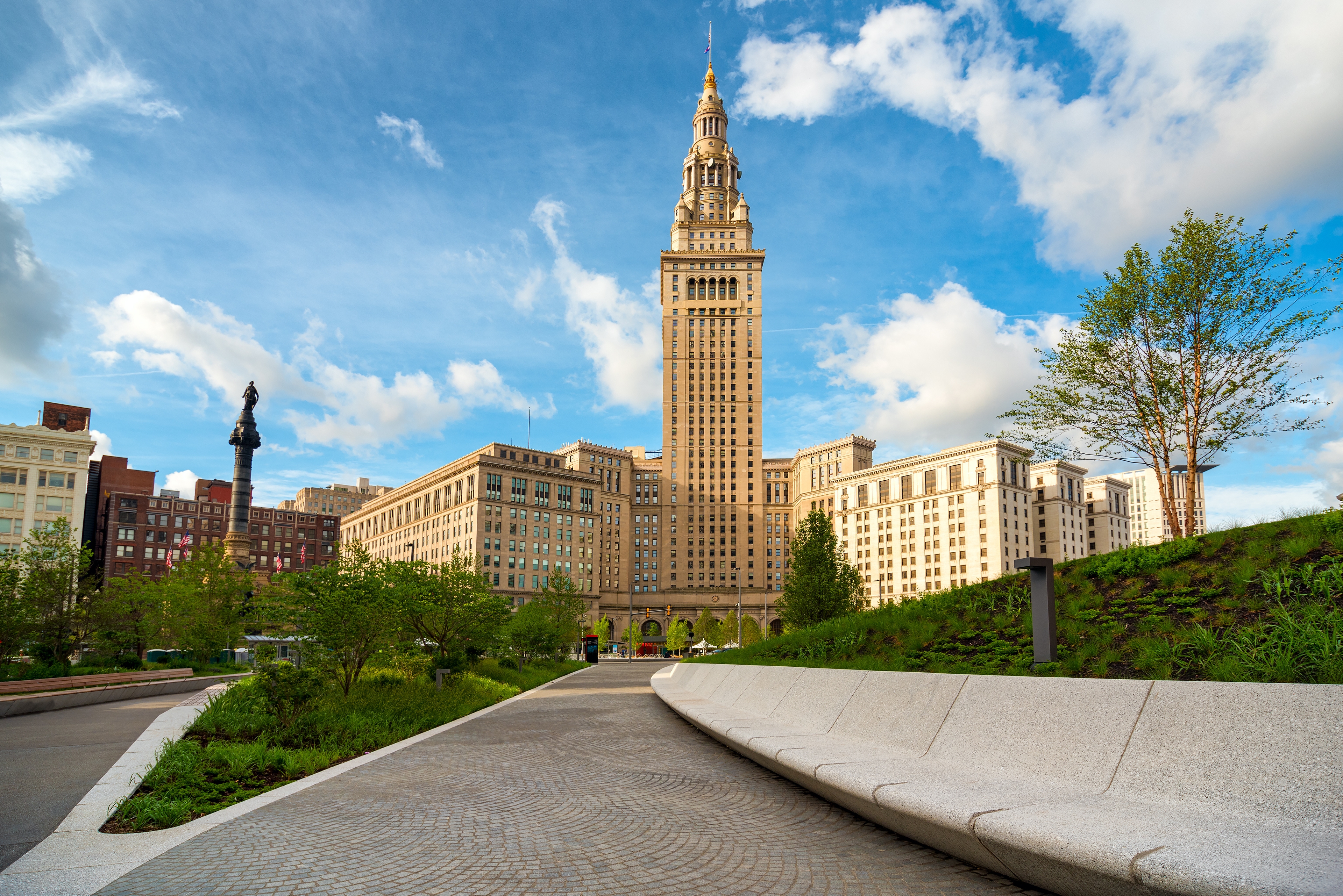 A sunny view of Cleveland's Terminal Tower, surrounded by historic buildings and greenery, with a curving pathway and modern seating in the foreground under a bright blue sky with scattered clouds.