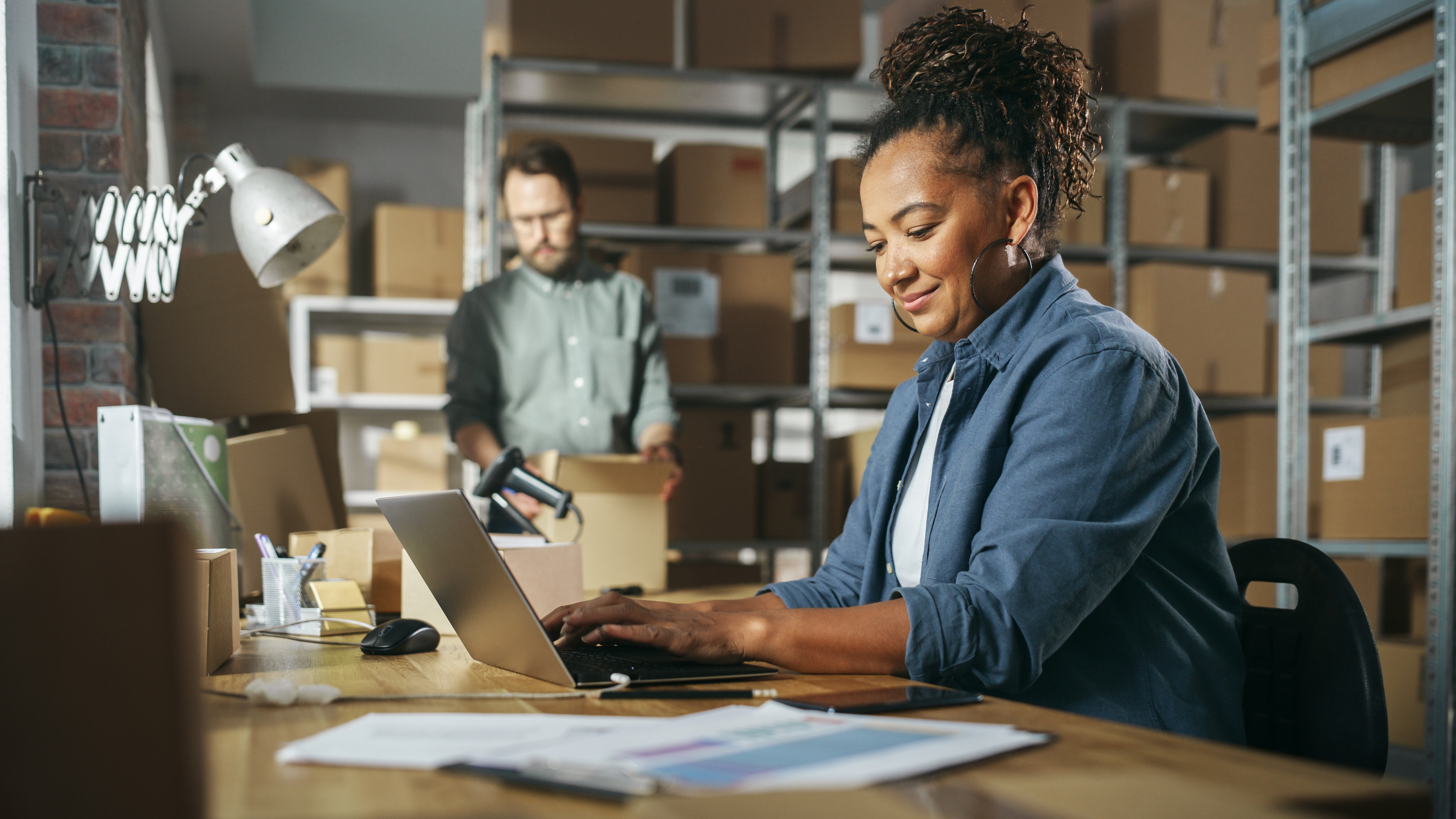 A woman smiling while working on a laptop in a warehouse, with a colleague organizing packages in the background. The scene suggests a busy and organized logistics or inventory management environment.