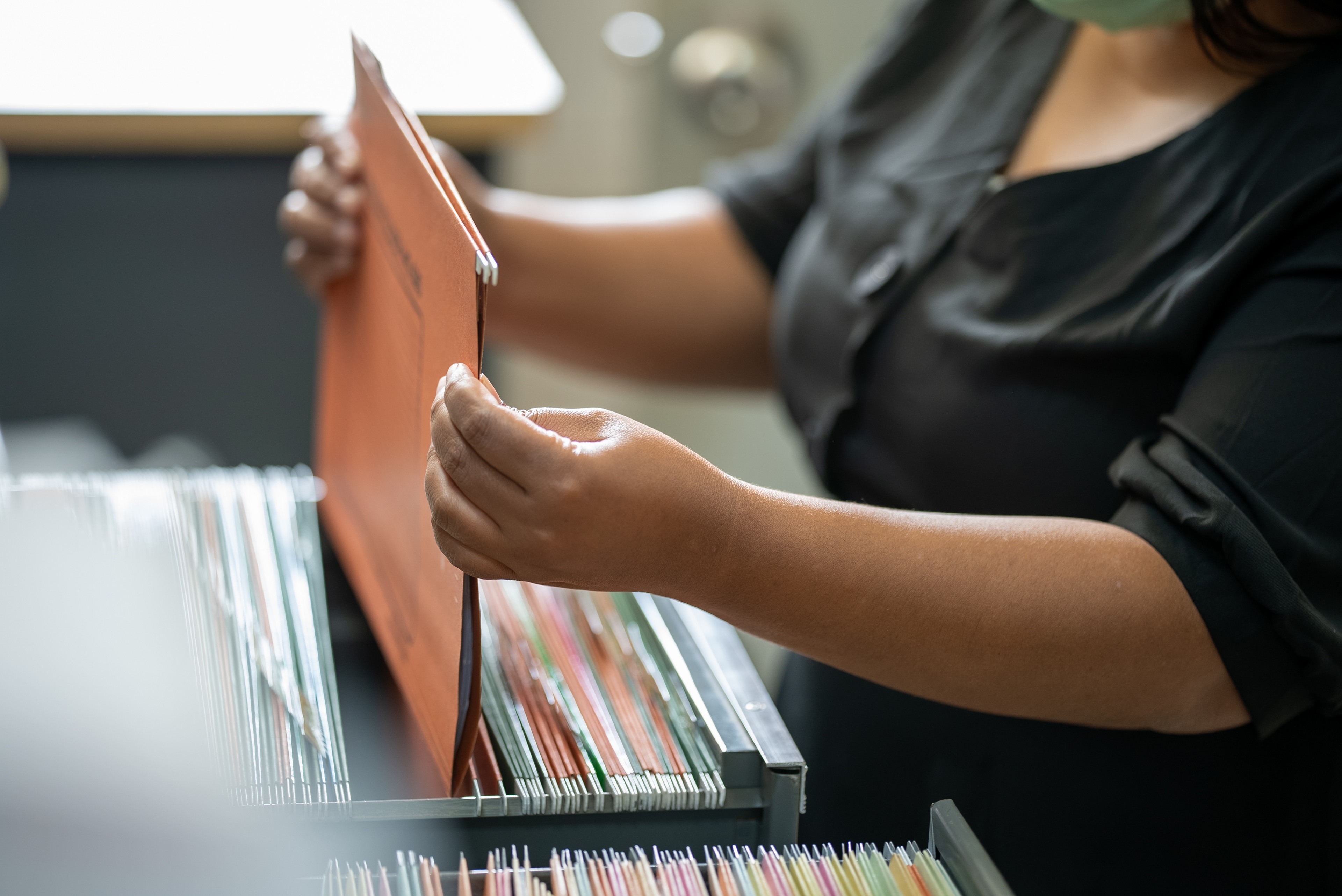 A person filing a document into a drawer filled with neatly organized folders. The individual is holding a brown file, carefully placing it into a file cabinet, indicating an organized filing or record-keeping process.
