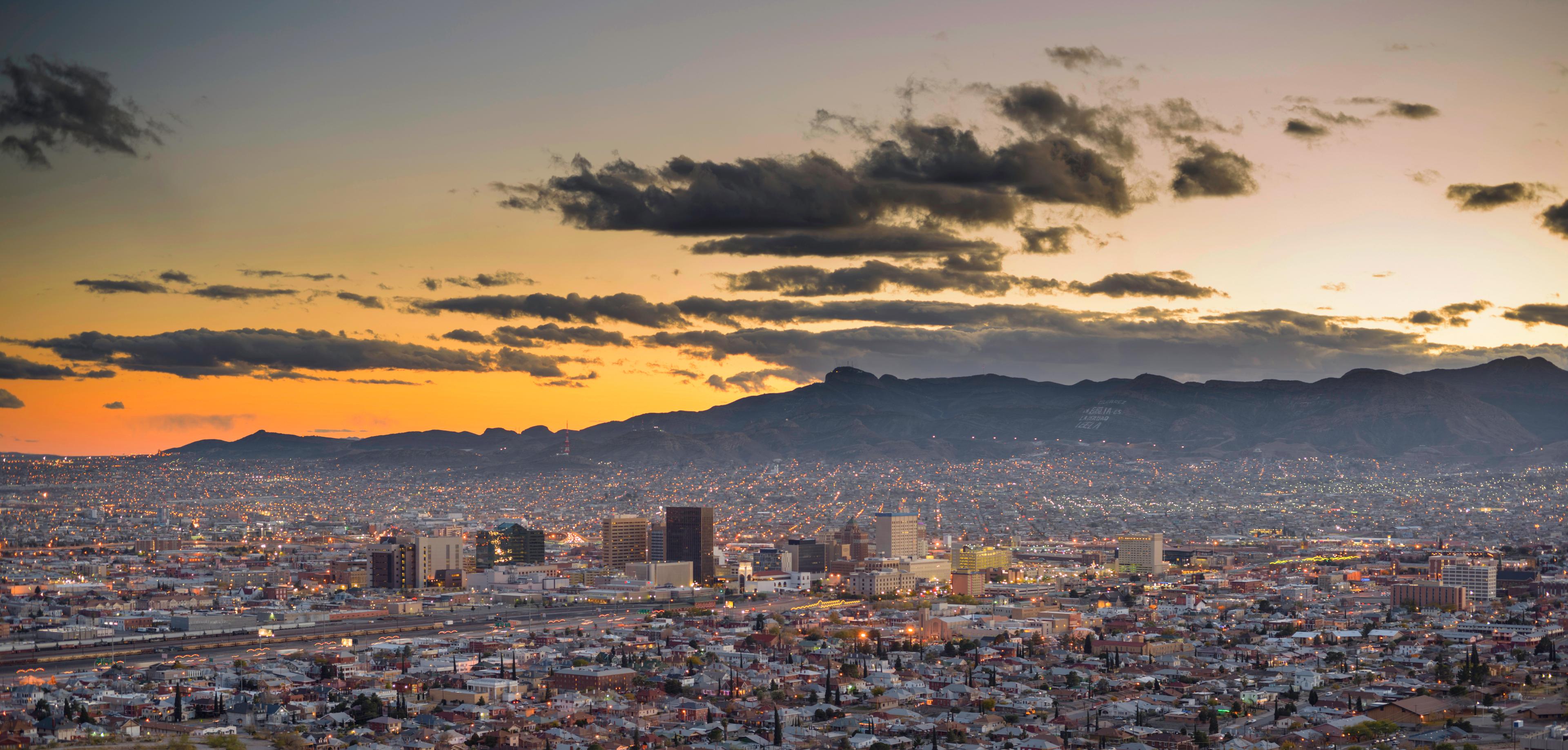 An evening view of El Paso, Texas, with its illuminated cityscape stretching to the horizon, framed by distant mountains and a vibrant orange and purple sunset. 