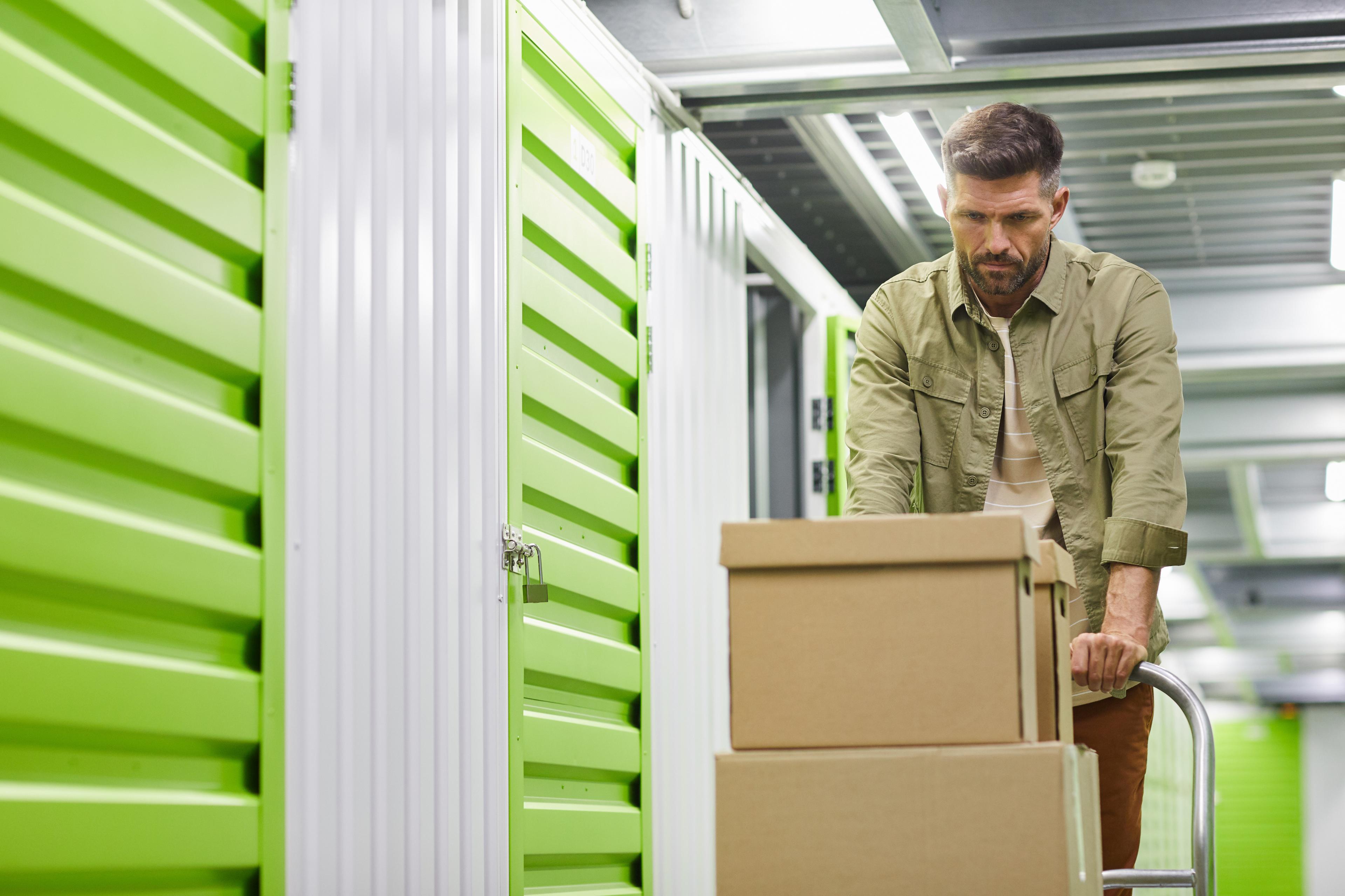 A man wearing a green jacket pushes a hand truck loaded with cardboard boxes down a hallway lined with bright green and white self-storage unit doors.