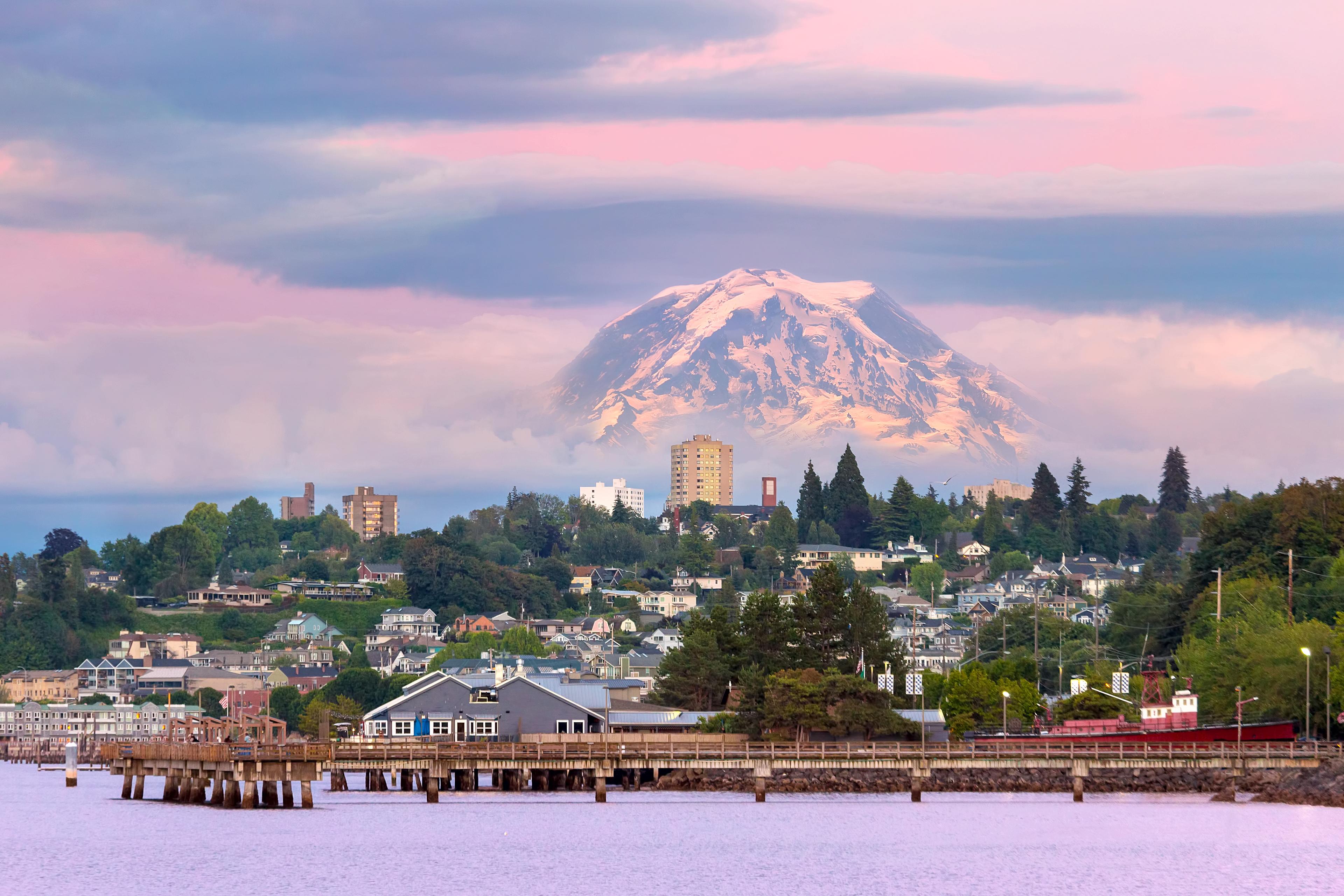 Dramatic view of Mount Rainier rising above the city of Tacoma, Washington, with a pastel sky at dusk and a wooden pier stretching over Puget Sound in the foreground.