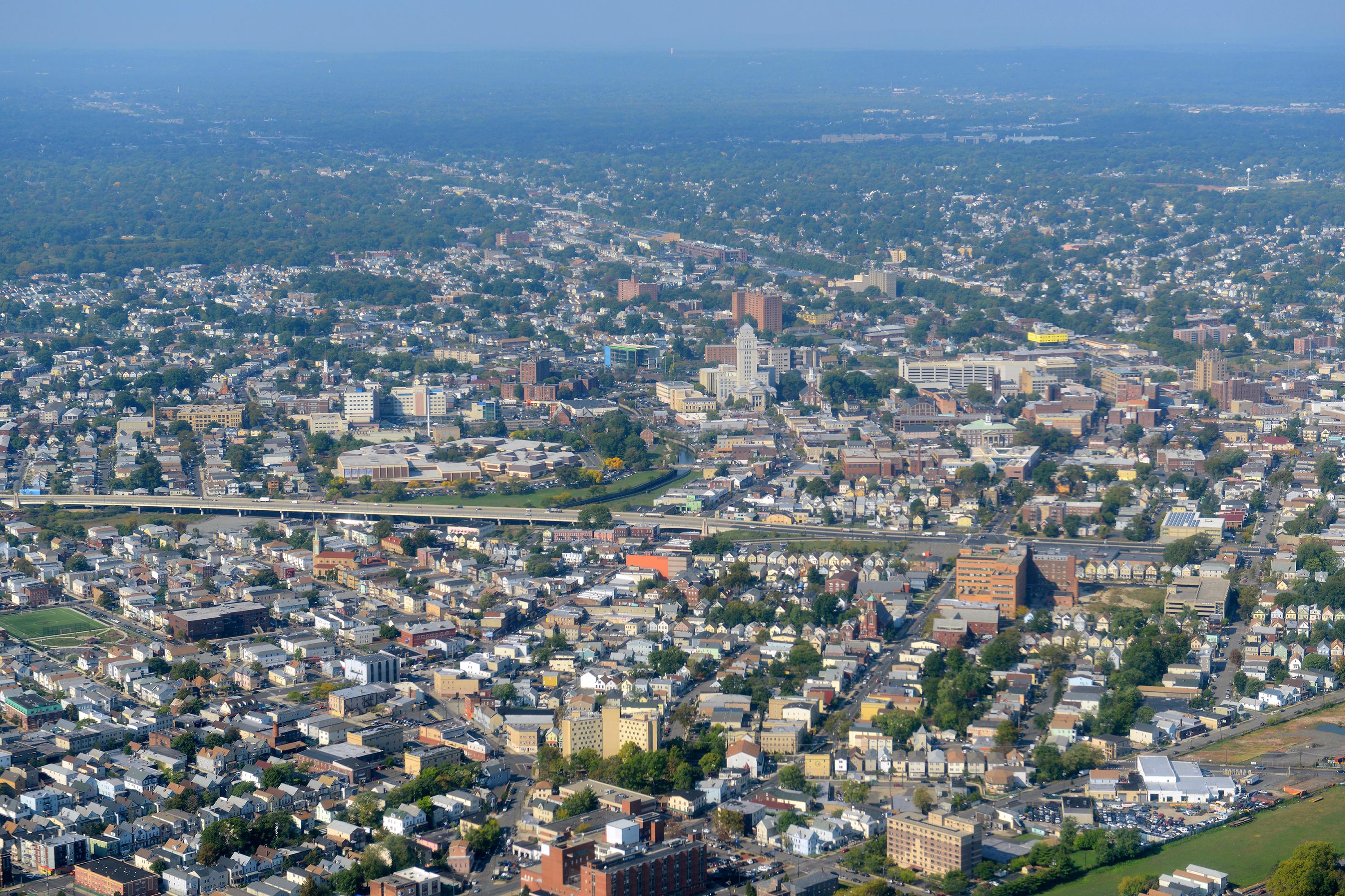 An aerial view of Elizabeth, New Jersey, showing a dense urban layout with residential neighborhoods, commercial buildings, and tree-lined streets. 