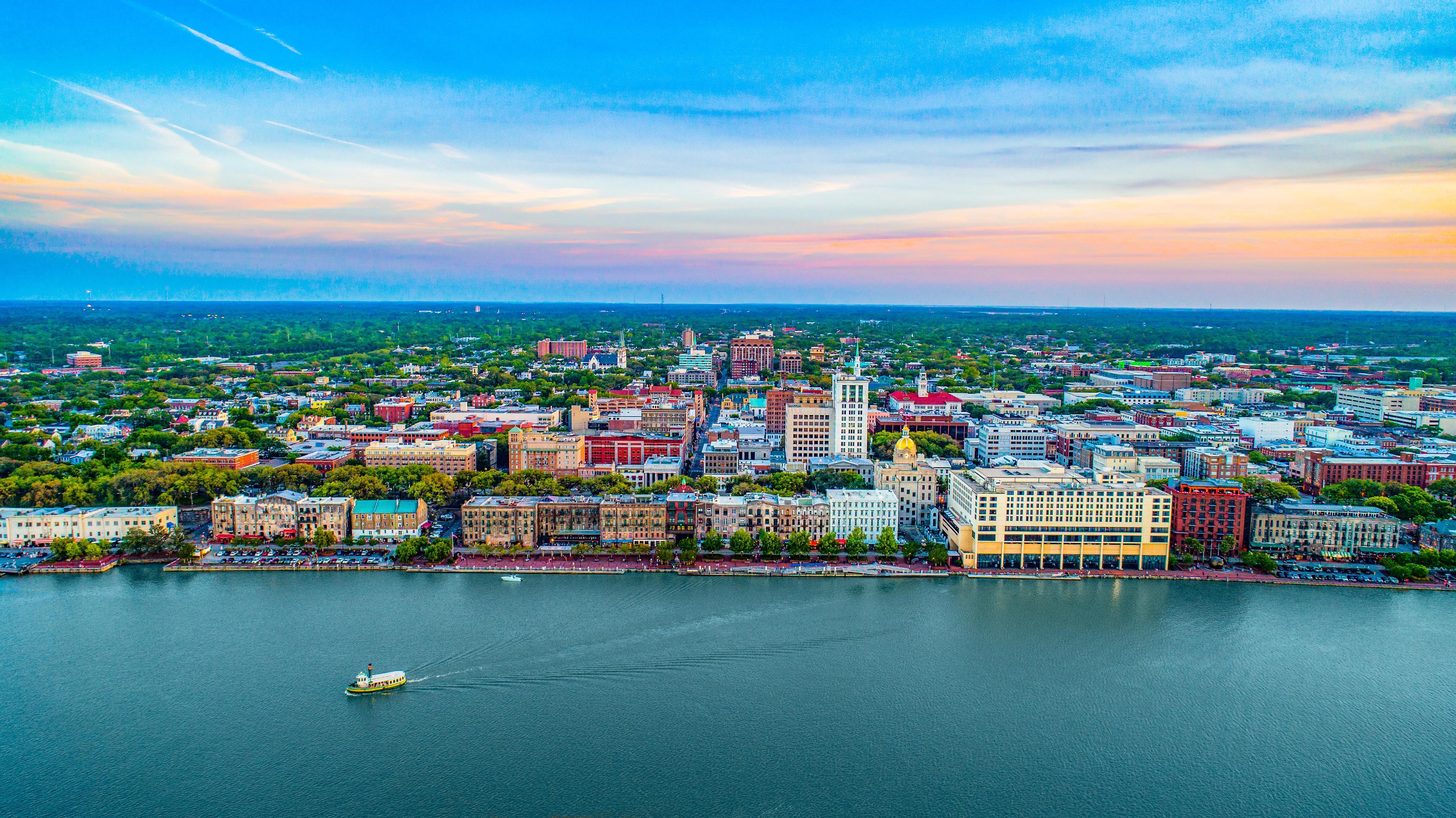 Colorful aerial view of Savannah, Georgia at sunset, with River Street buildings lining the Savannah River and a ferryboat cruising across calm waters beneath a pastel sky.