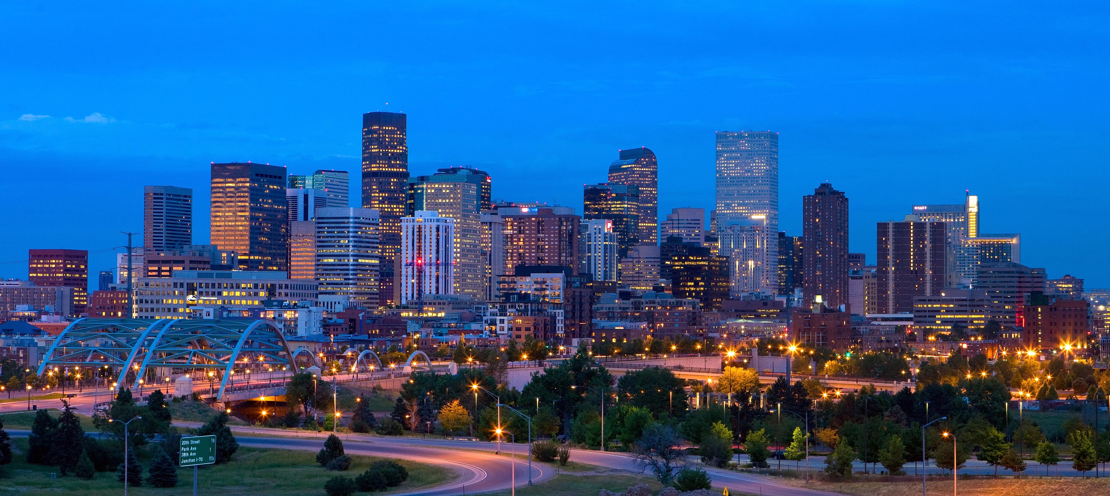 A wide-angle twilight view of downtown Denver features illuminated skyscrapers and a modern arched bridge, with glowing city lights against a vivid blue sky.