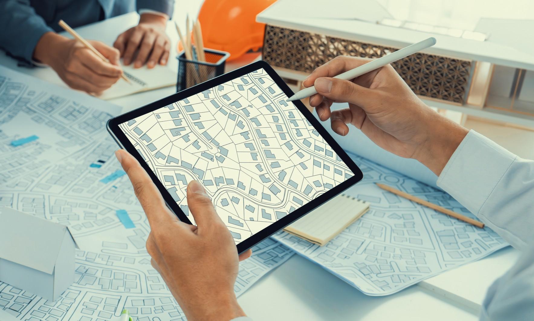 Close-up of an real estate planner using a digital tablet with a zoning map, surrounded by real estate documents, a model building, and drafting tools during a planning meeting.